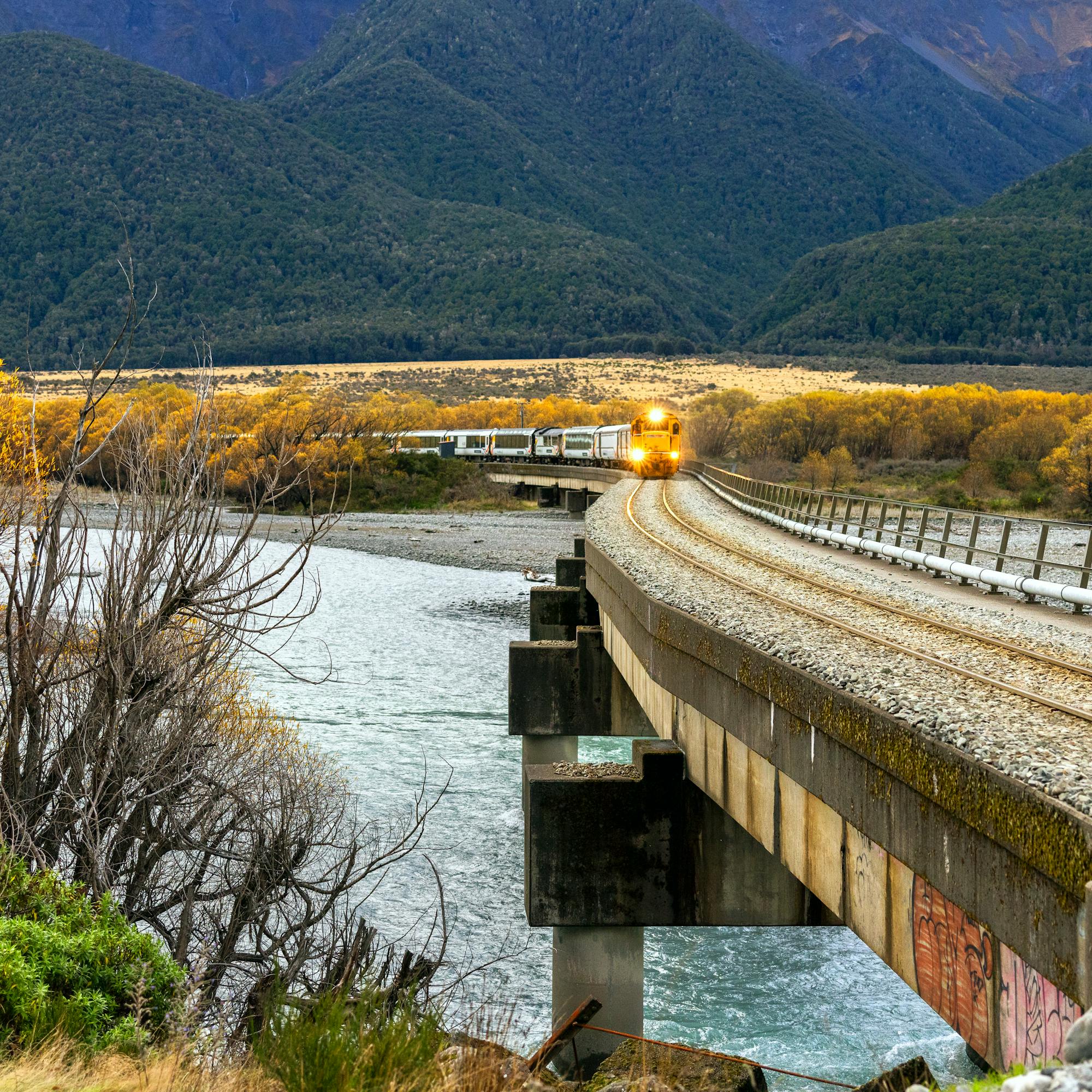 Milky turquoise river curves past a stone embankment and forested hillside, with low clouds over distant peaks.
