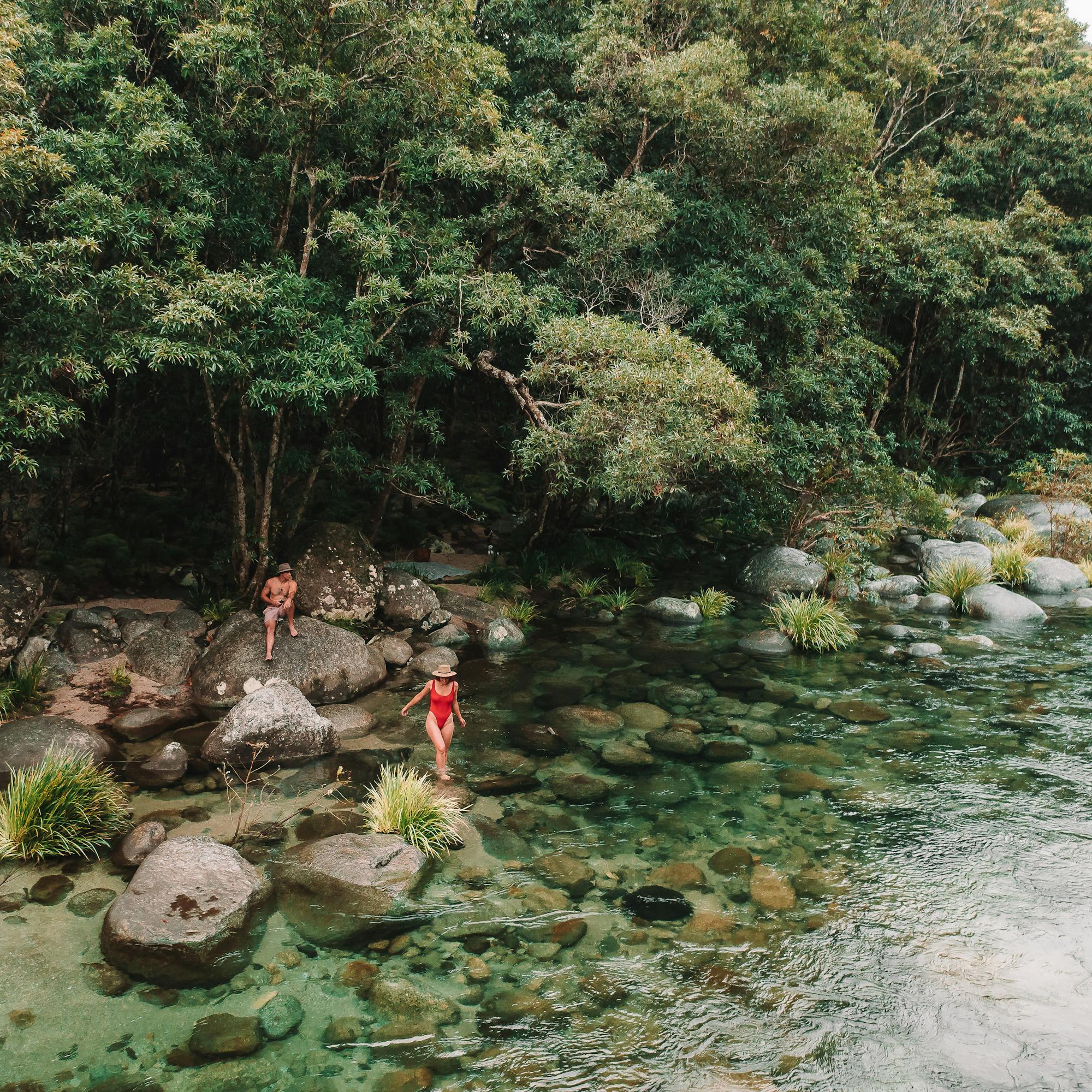 Two guests wade in a clear rainforest river, mossy boulders and dense green canopy edging the quiet waterway.