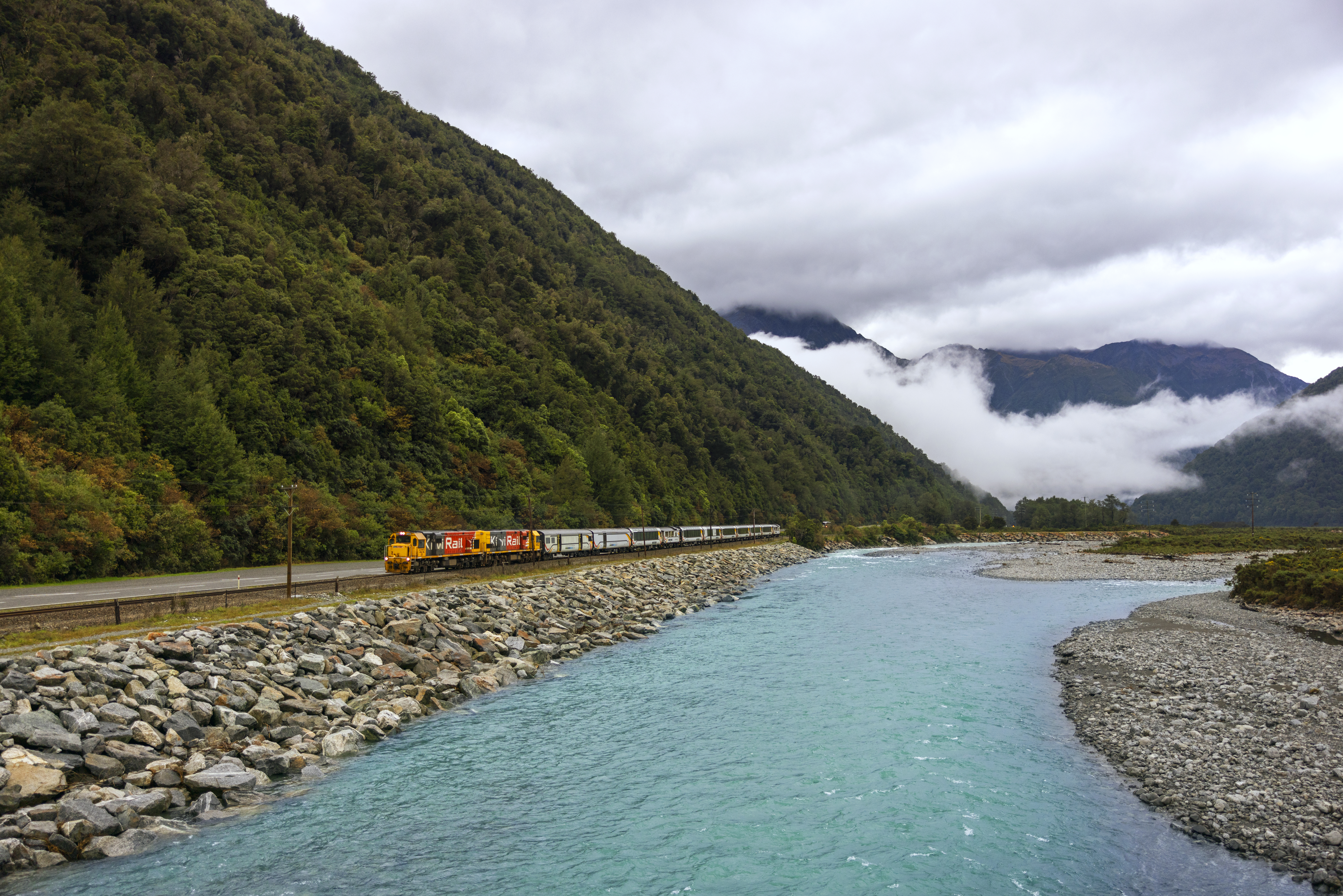 KiwiRail train runs beside a turquoise river, skirting a rocky embankment below forested mountains and mist.