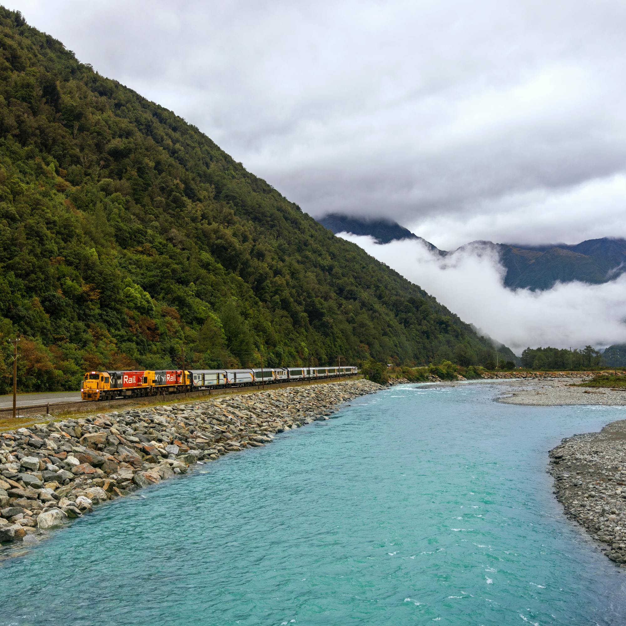 KiwiRail train runs beside a turquoise river, skirting a rocky embankment below forested mountains and mist.