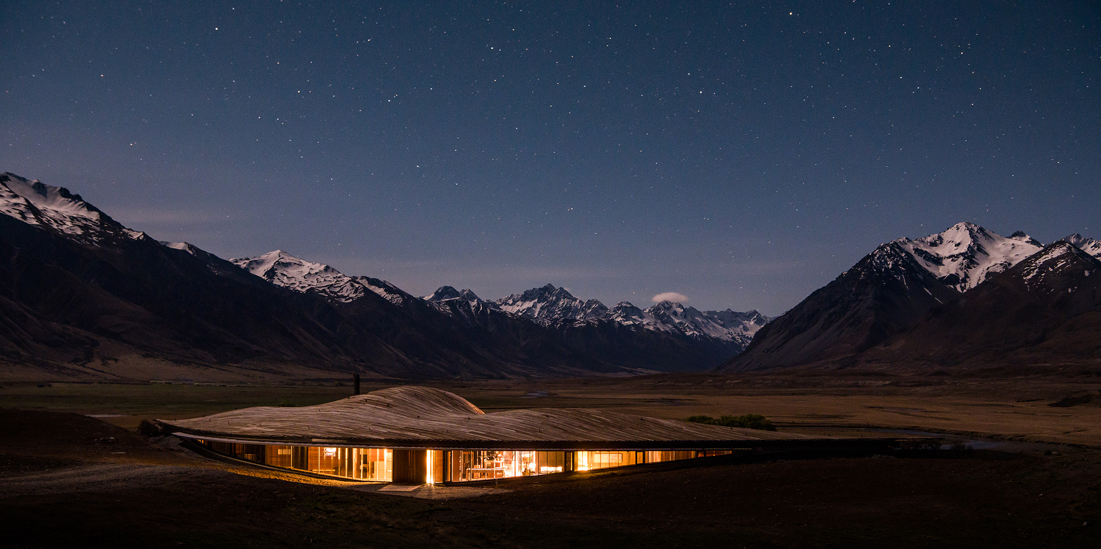 Lodge lights glow quietly in a mountain valley under a starry sky, with dark peaks and snowy ridges beyond.