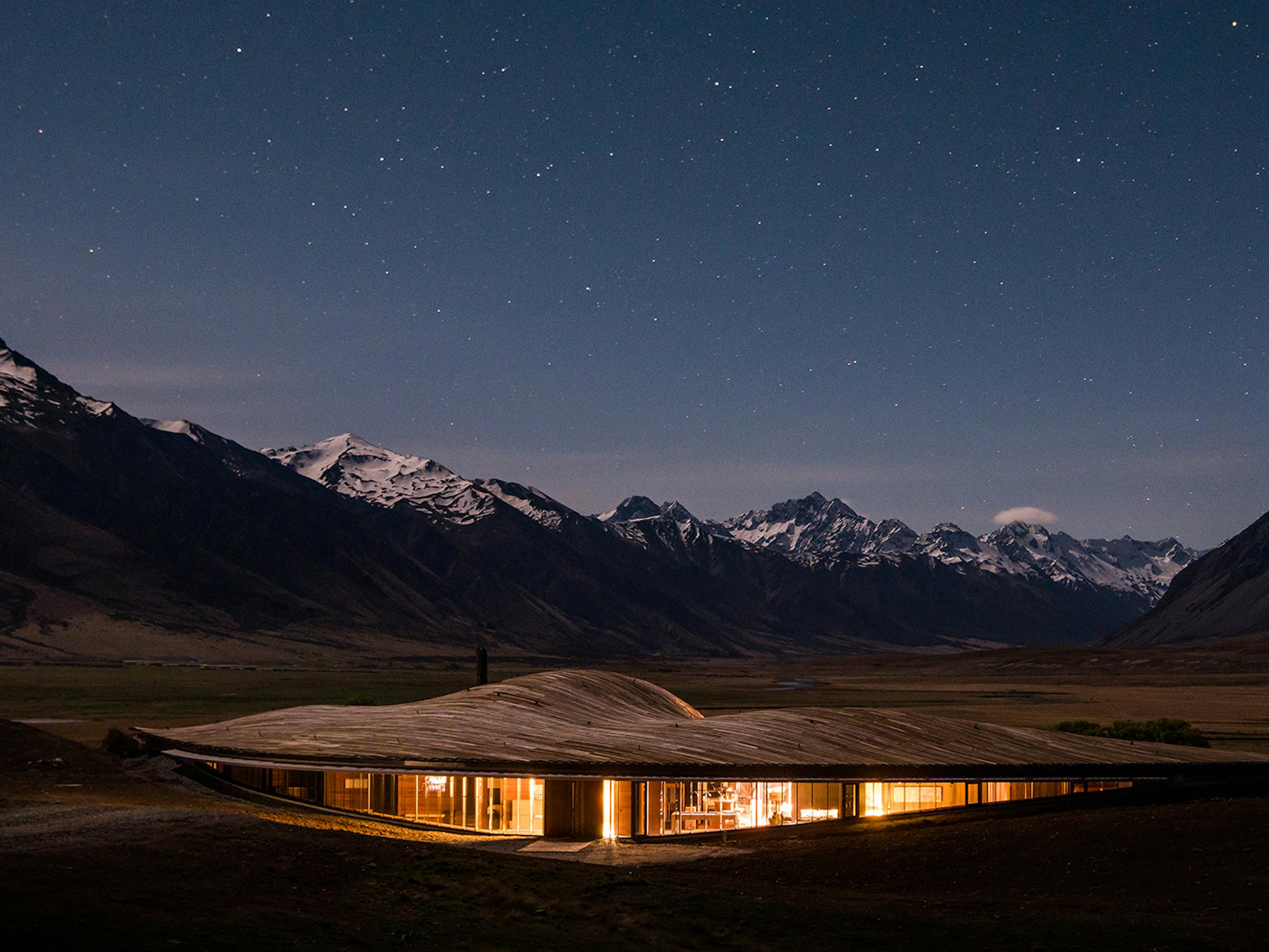 Lodge lights glow quietly in a mountain valley under a starry sky, with dark peaks and snowy ridges beyond.