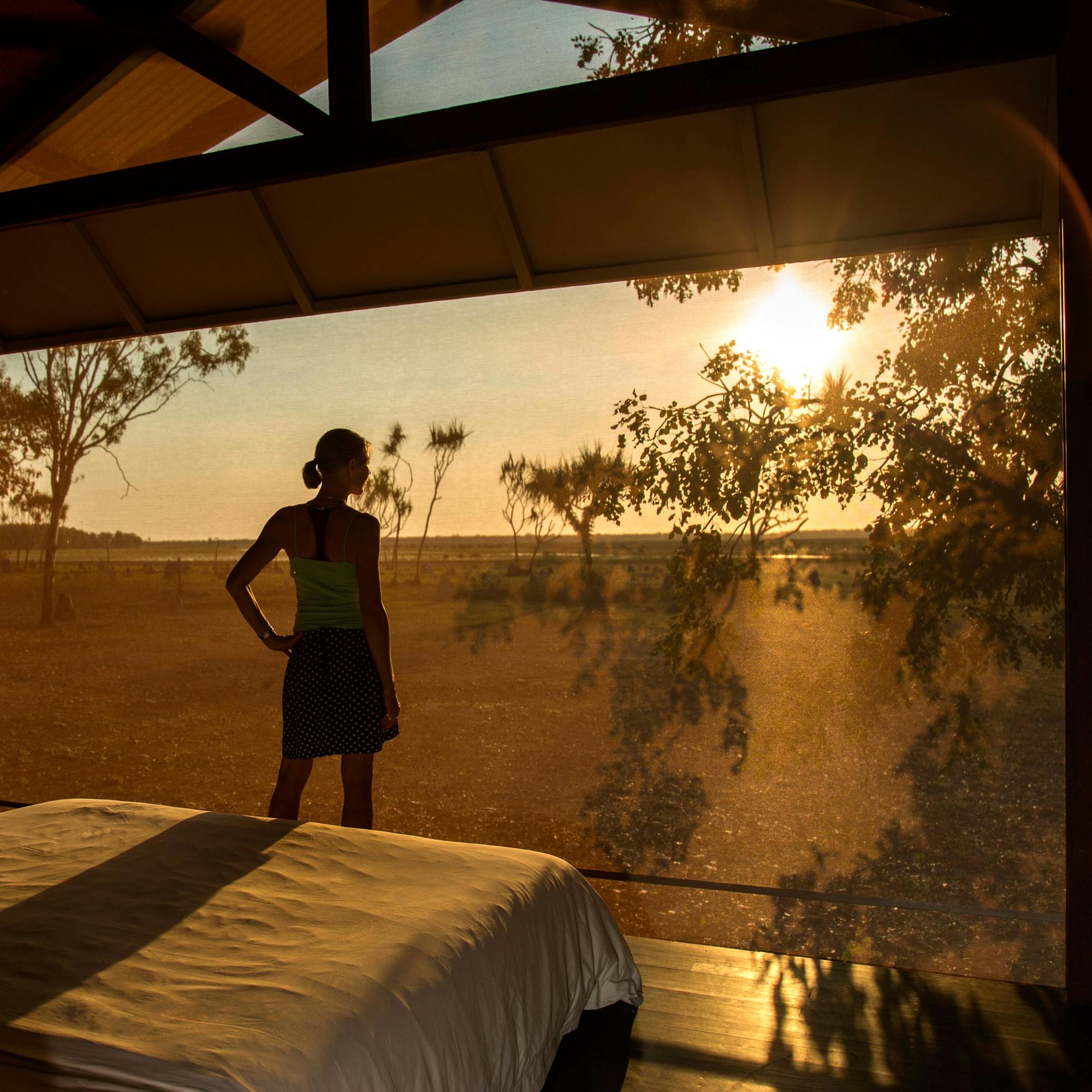 A guest stands inside a canvas tent beside the bed, looking out toward glowing sunset light over open wetlands.