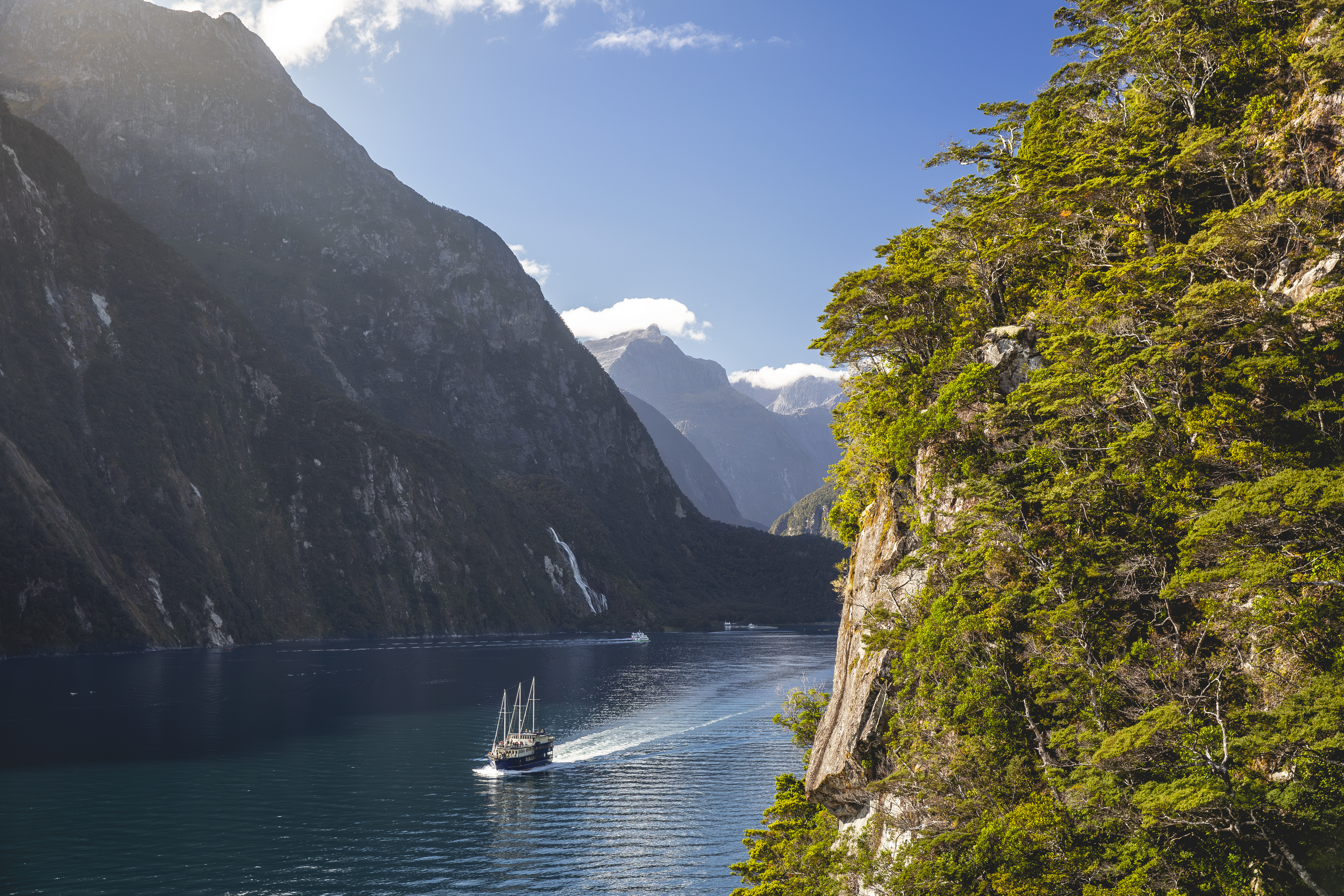 A small boat travels Milford Sound beneath towering dark cliffs, with sunlit water and green forest hanging to the edge.