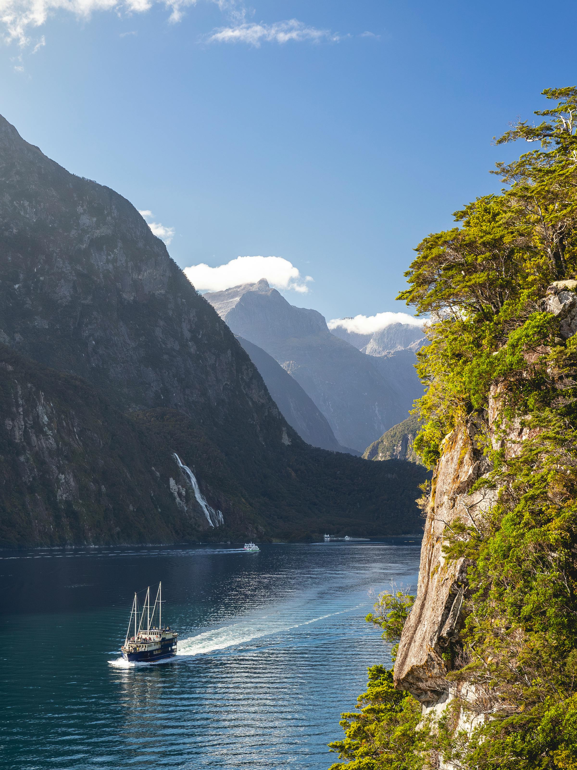 A small boat travels Milford Sound beneath towering dark cliffs, with sunlit water and green forest hanging to the edge.