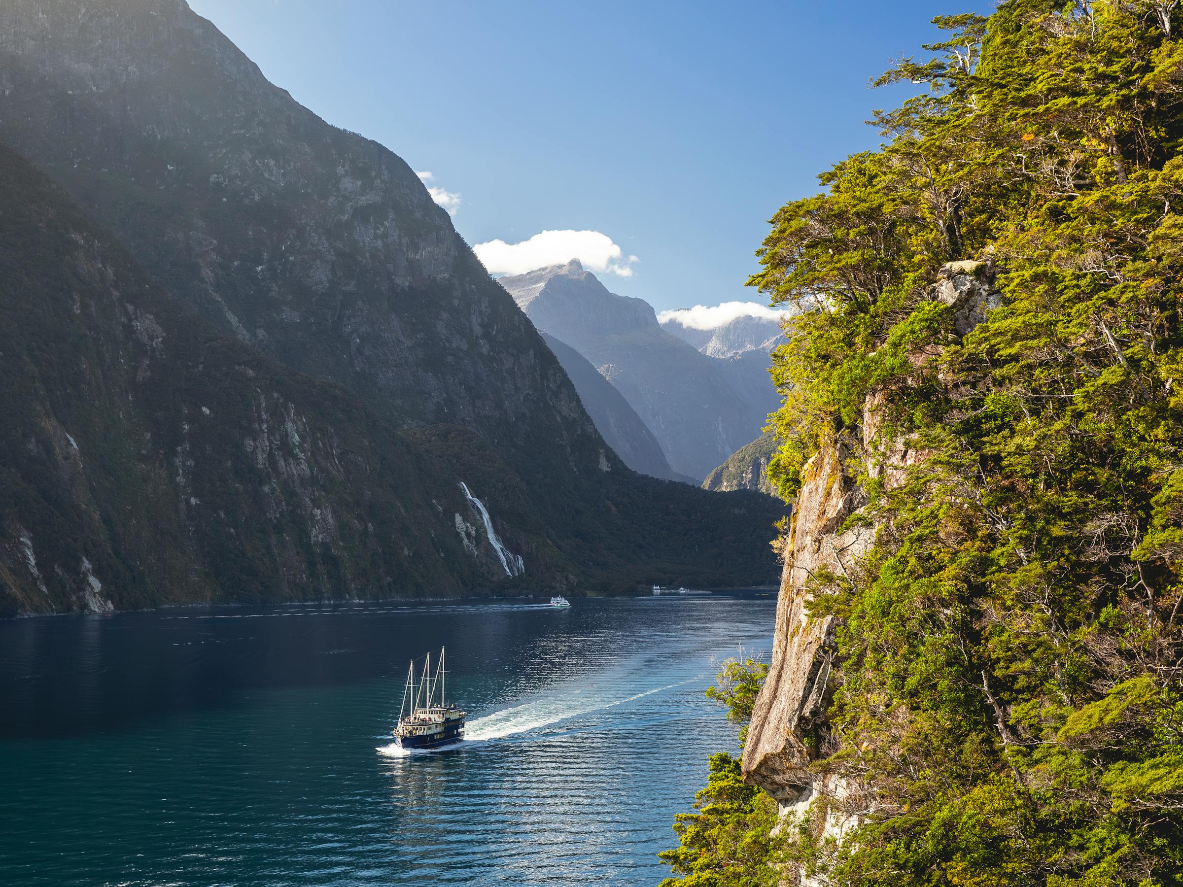 A small boat travels Milford Sound beneath towering dark cliffs, with sunlit water and green forest hanging to the edge.