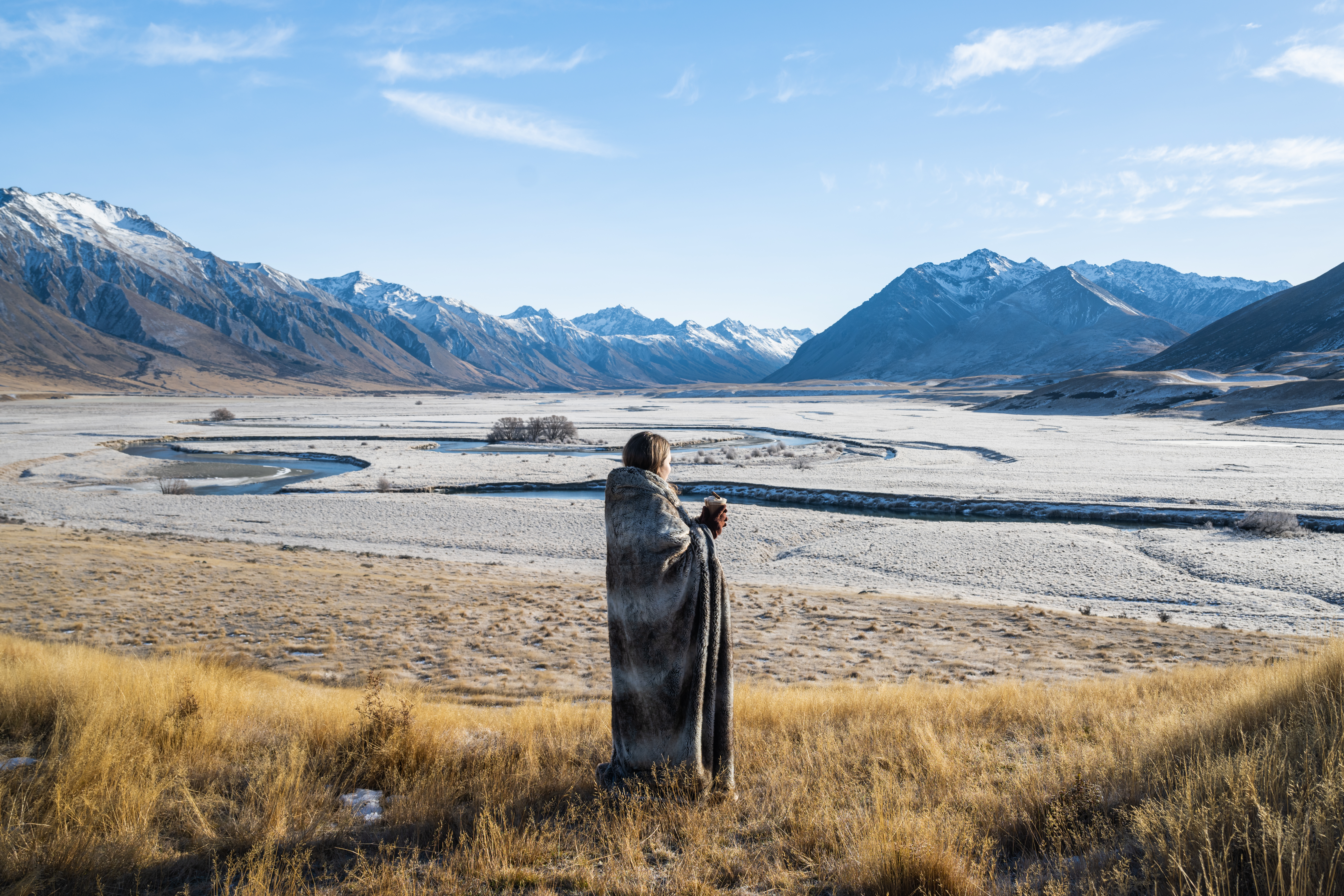 A lone traveler stands at a viewpoint over a braided river plain, with golden tussock and snowy mountains ahead.