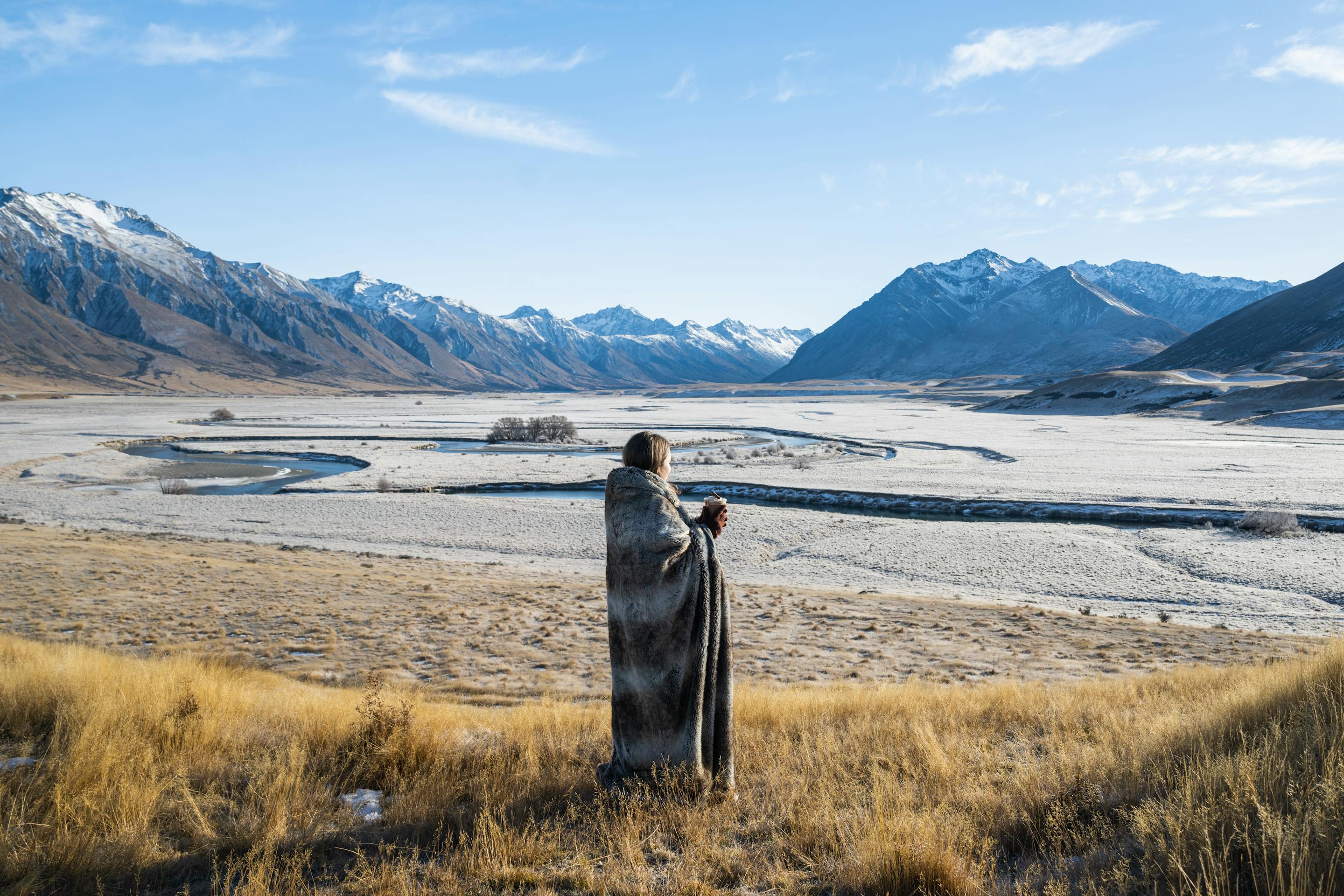A lone traveler stands at a viewpoint over a braided river plain, with golden tussock and snowy mountains ahead.