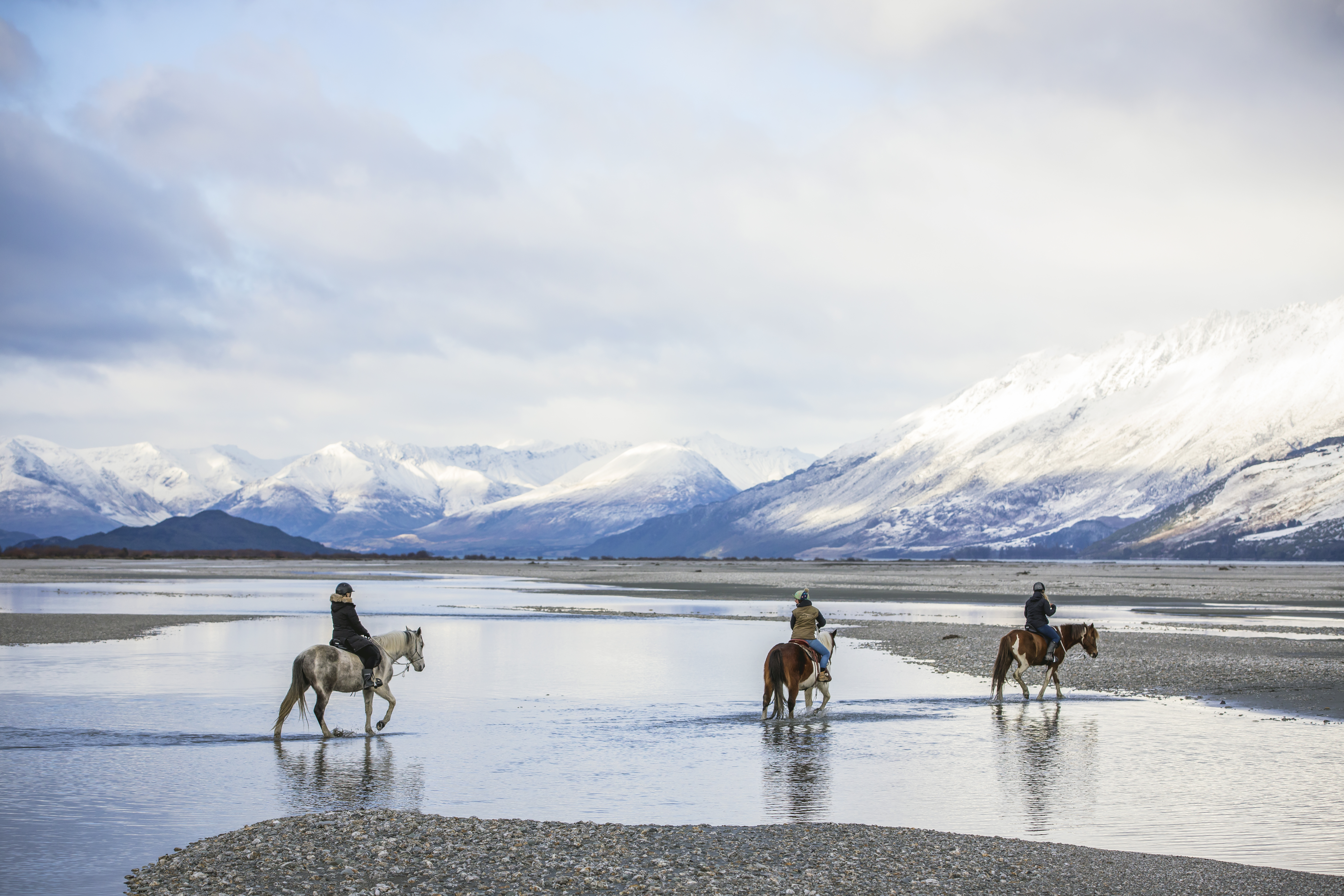 Three riders cross a shallow braided river, their horses reflected in calm water with snowy peaks beyond.