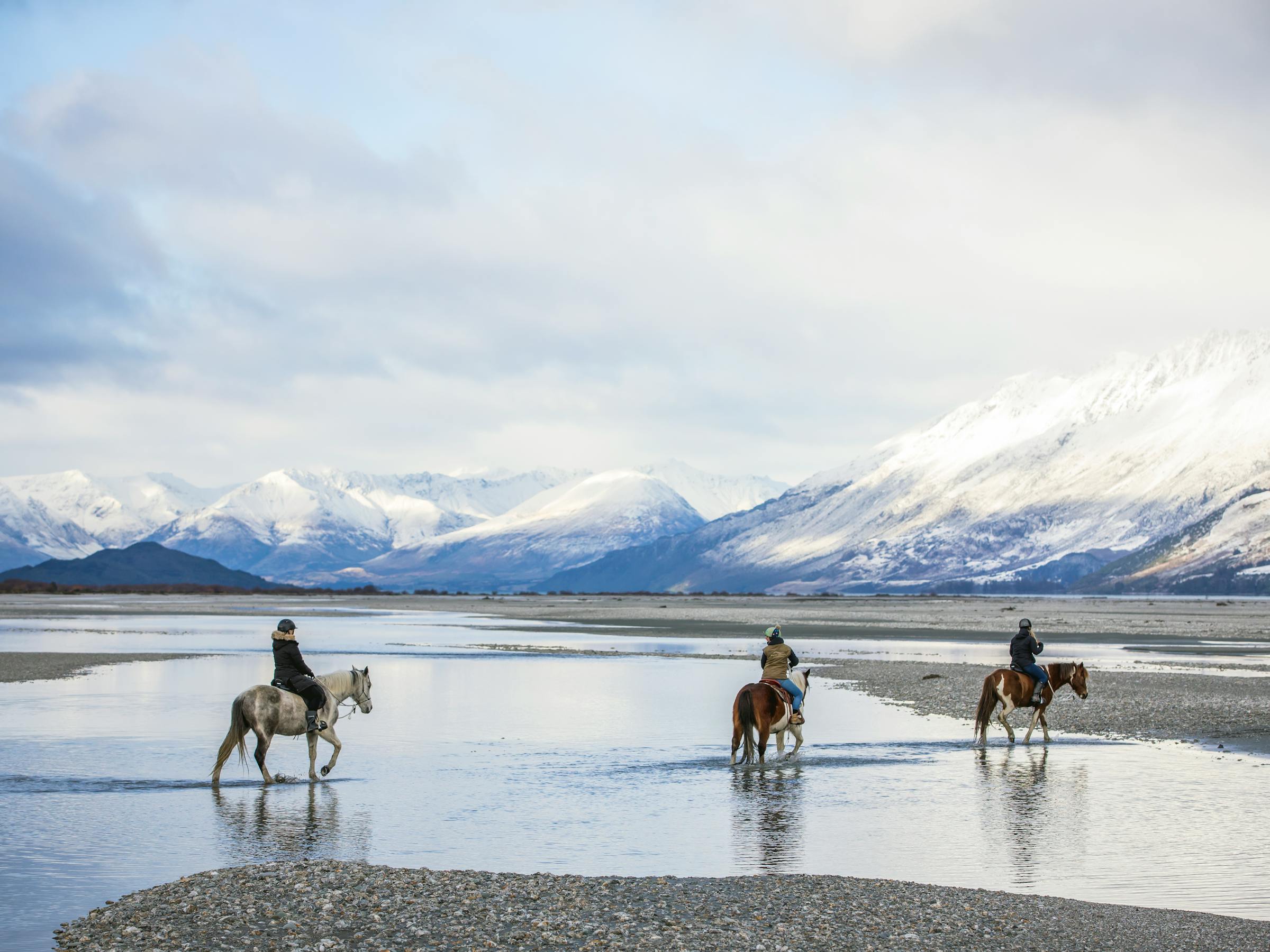 Three riders cross a shallow braided river, their horses reflected in calm water with snowy peaks beyond.