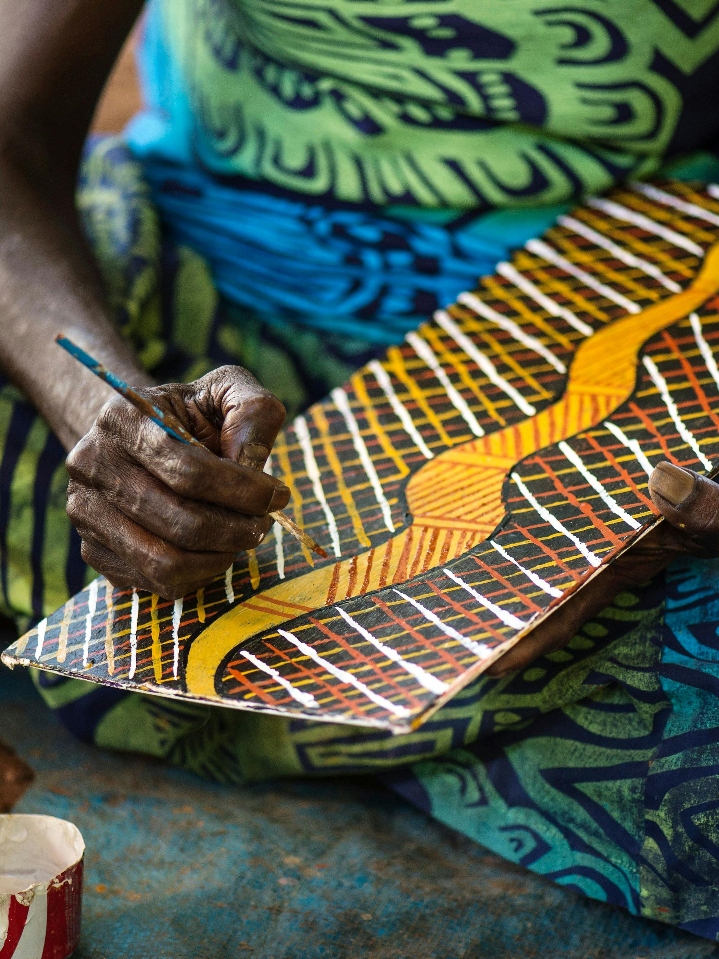 Artist paints a patterned wooden panel with fine lines and ochre tones, holding a brush above small paint pots.