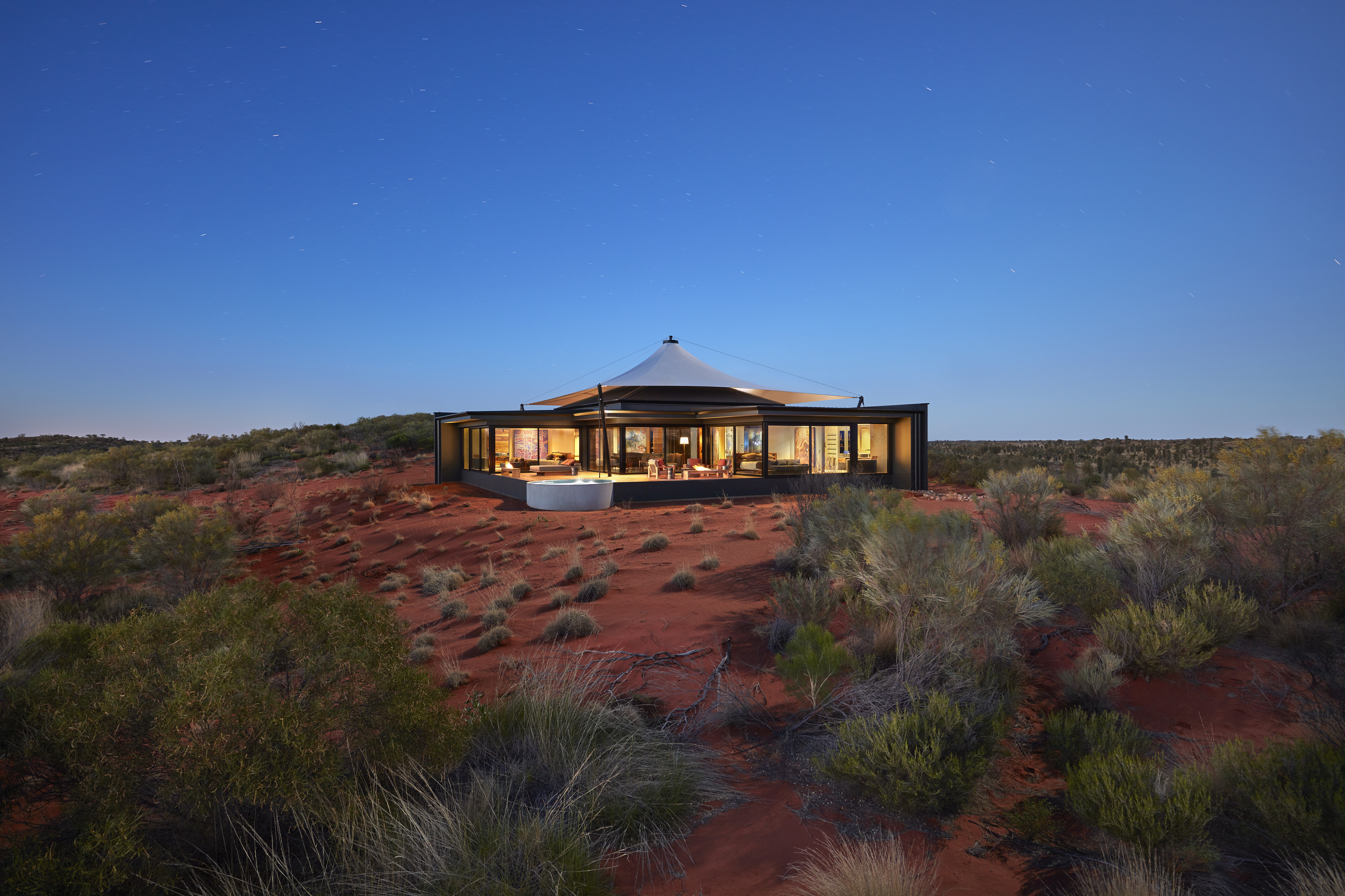 A low pavilion glows at sunset on red desert sand, surrounded by spinifex and sparse shrubs beneath blue sky.