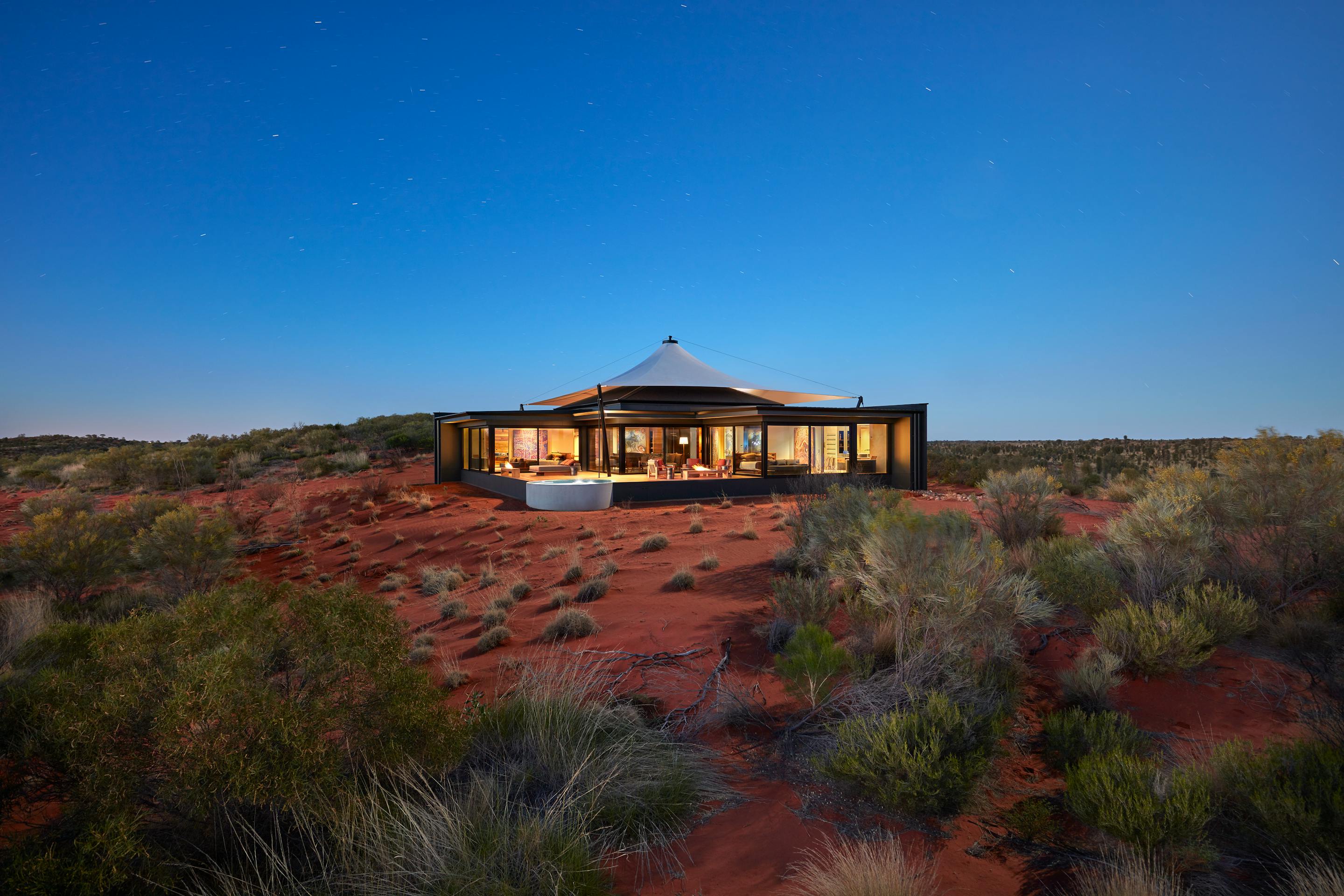 A low pavilion glows at sunset on red desert sand, surrounded by spinifex and sparse shrubs beneath blue sky.