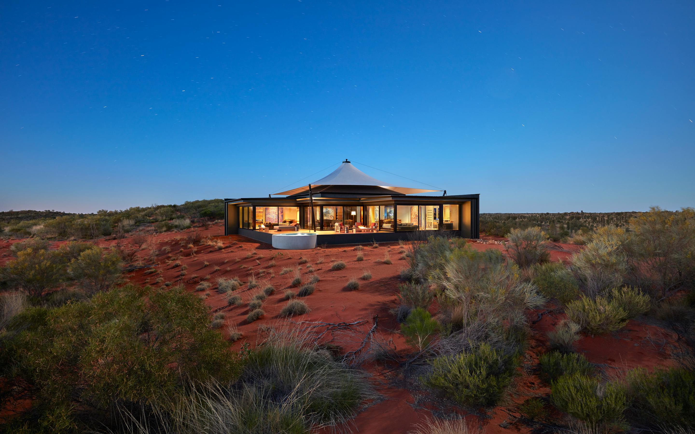 A low pavilion glows at sunset on red desert sand, surrounded by spinifex and sparse shrubs beneath blue sky.
