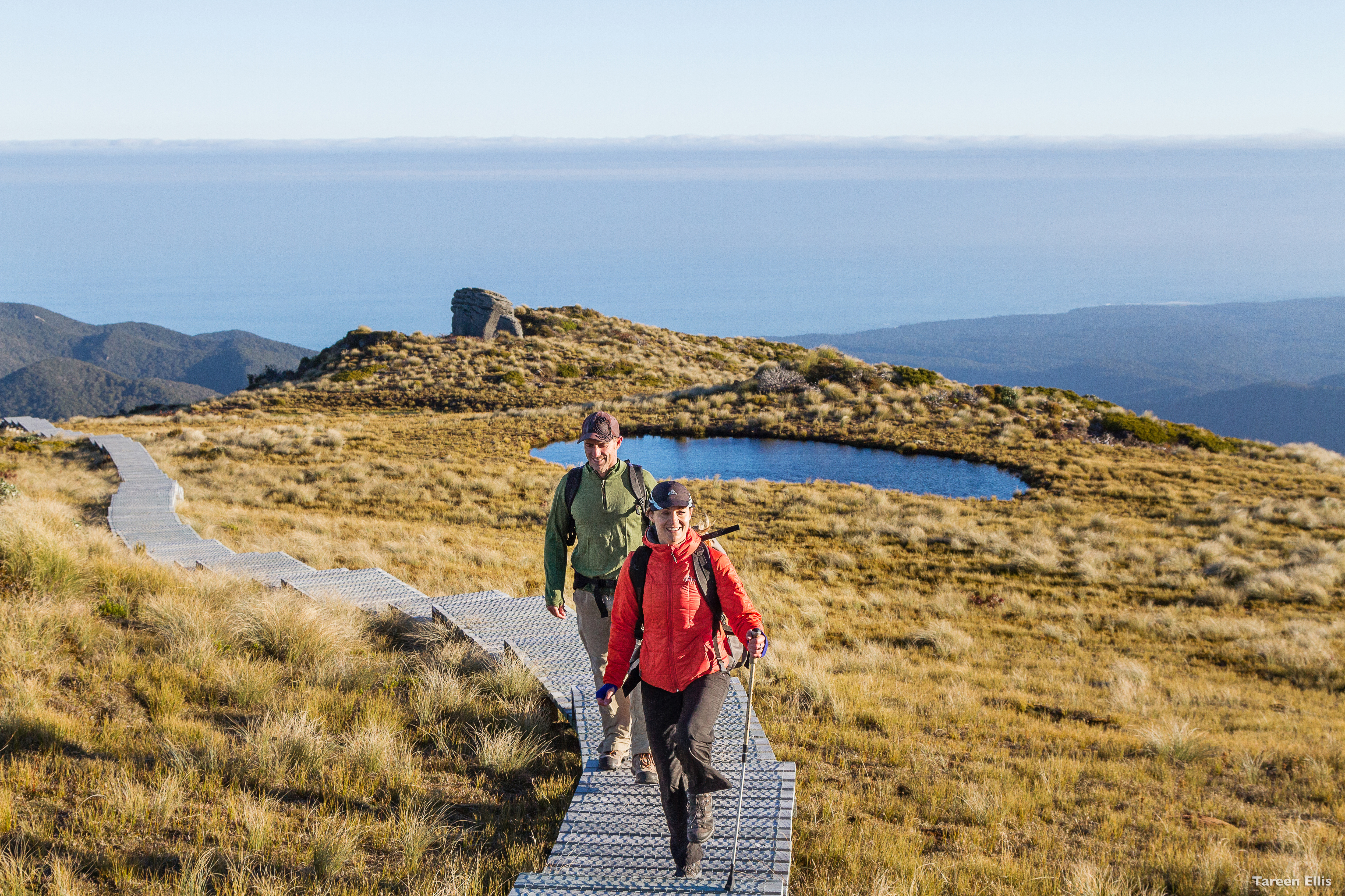 Two hikers walk a boardwalk across golden tussock toward small alpine tarns, with distant sea on the horizon.