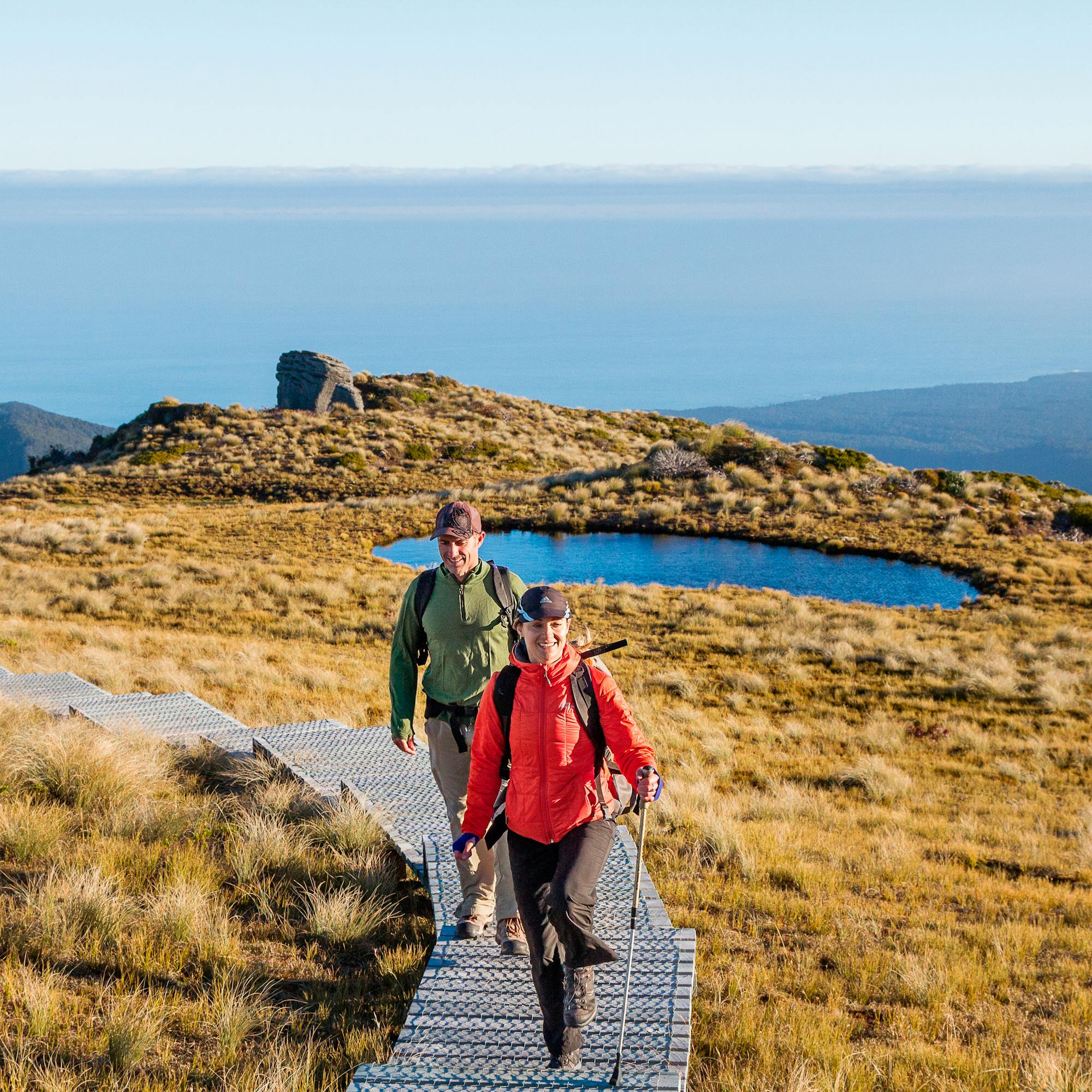 Two hikers walk a boardwalk across golden tussock toward small alpine tarns, with distant sea on the horizon.
