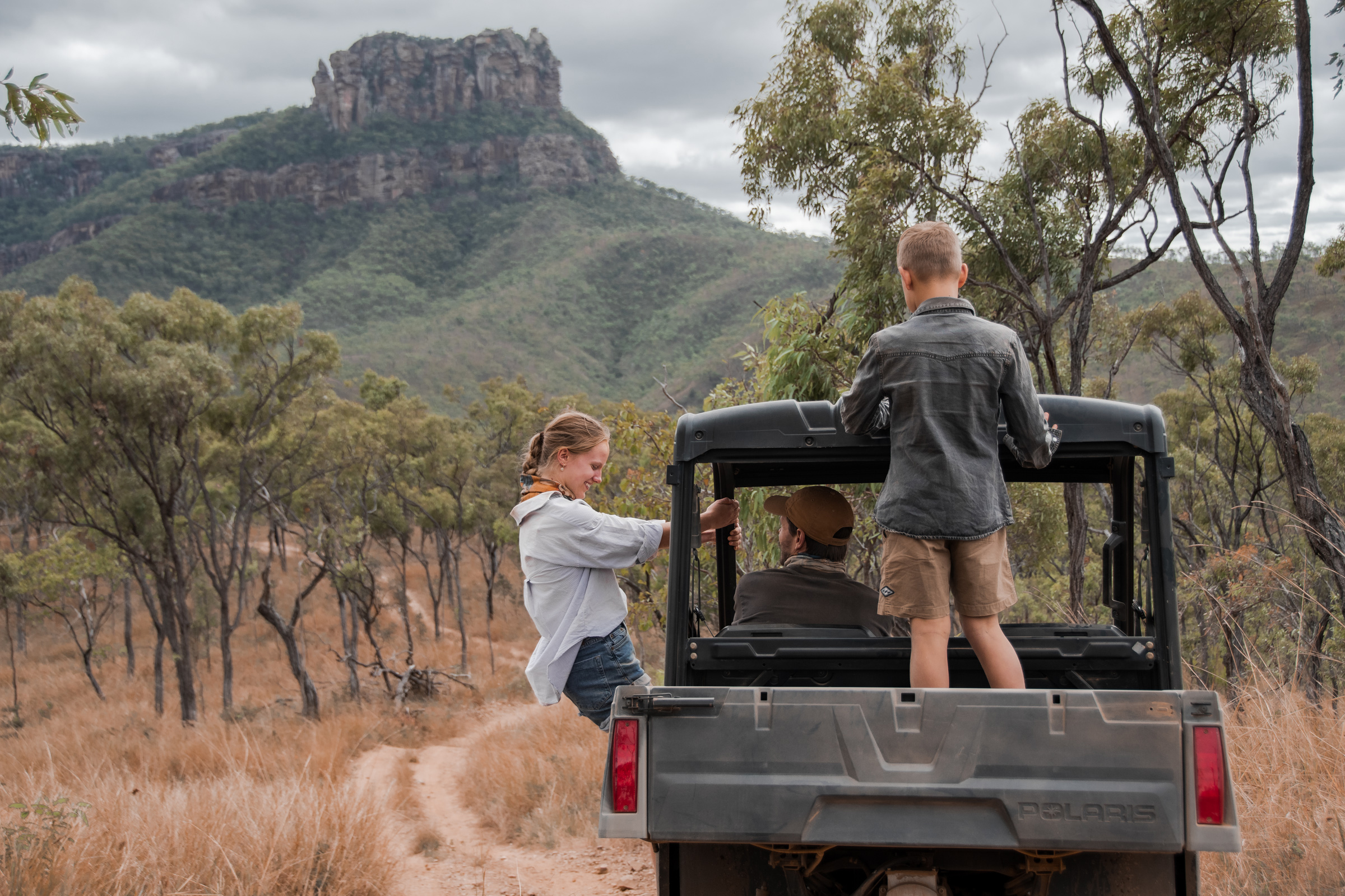 A family stands on an open safari vehicle, watching the rugged outback landscape and mountains ahead on the track.