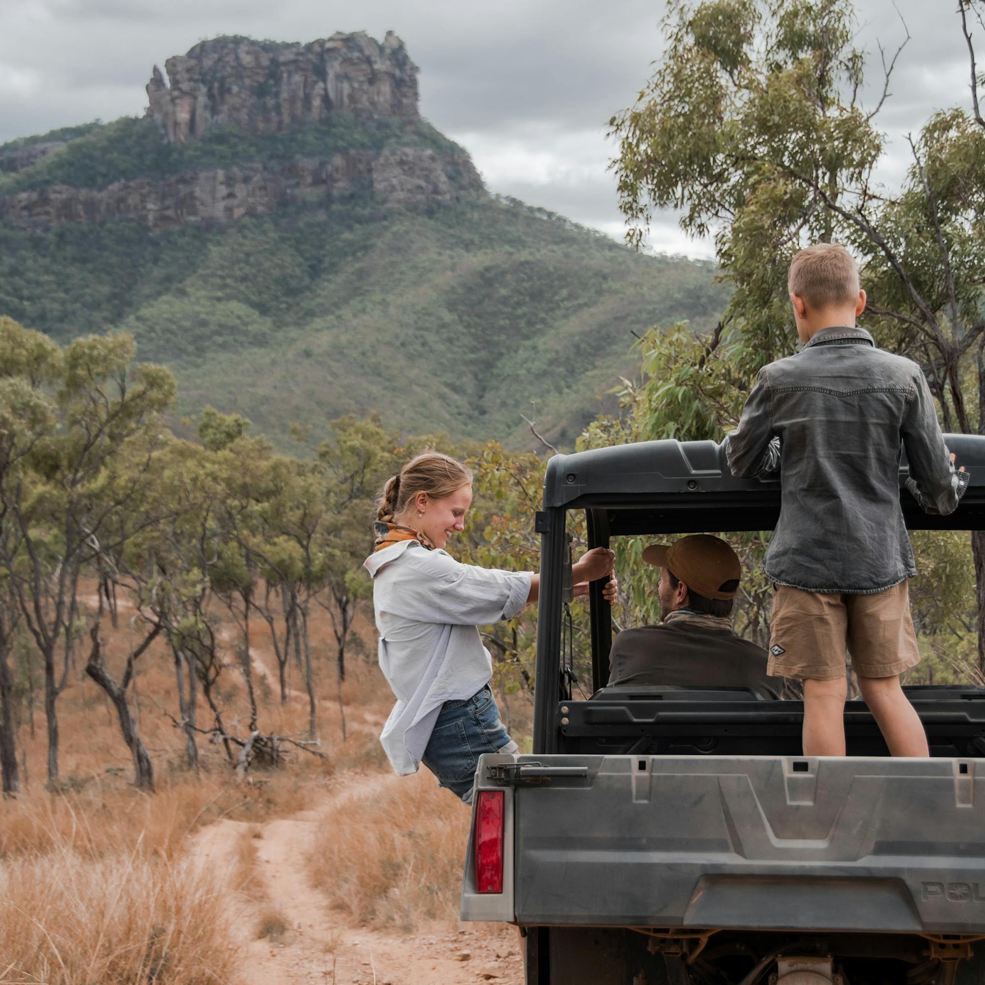 A family stands on an open safari vehicle, watching the rugged outback landscape and mountains ahead on the track.