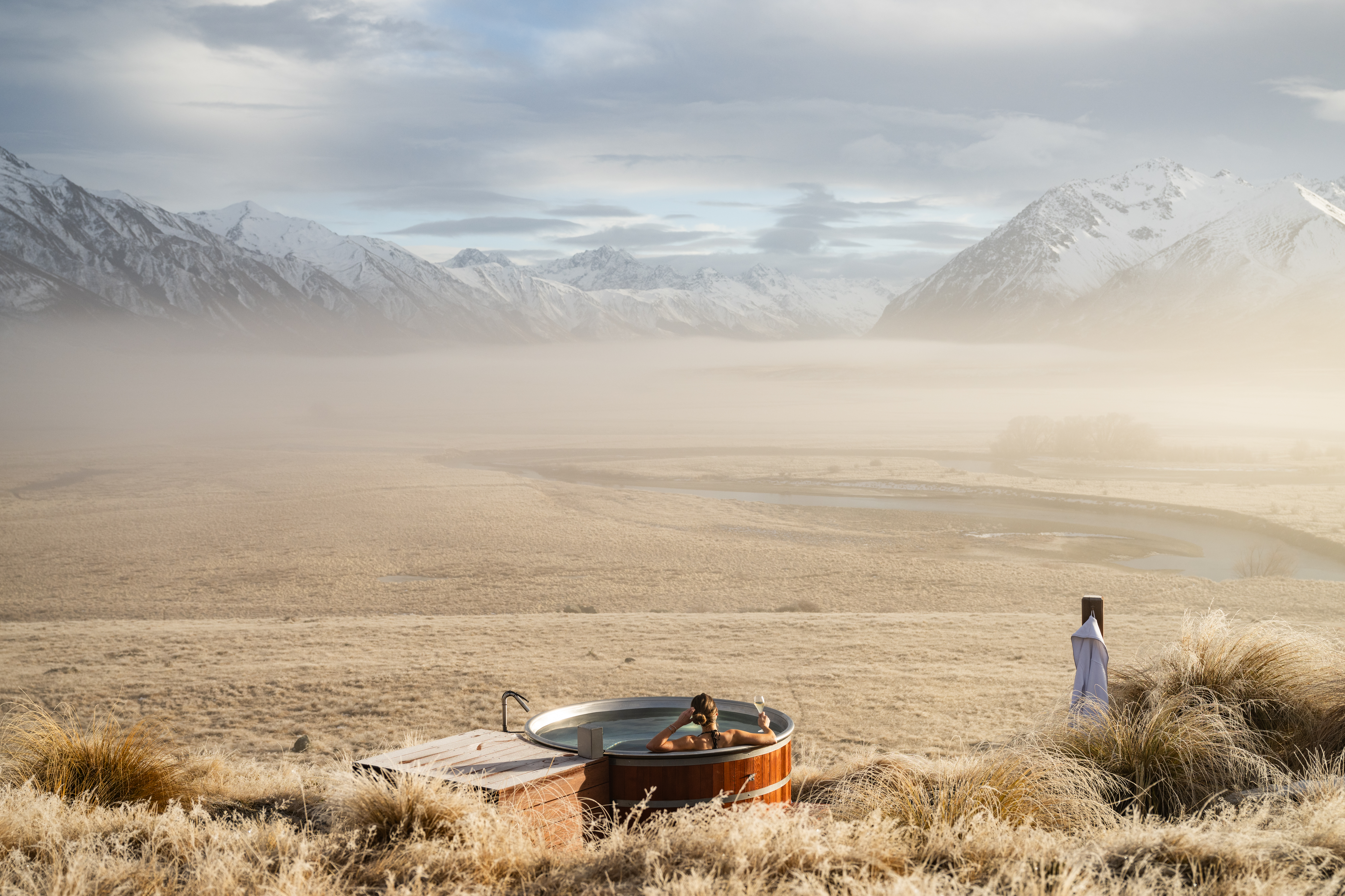 An outdoor hot tub sits in dry tussock grass as a lone guest looks across a misty valley toward rugged peaks.