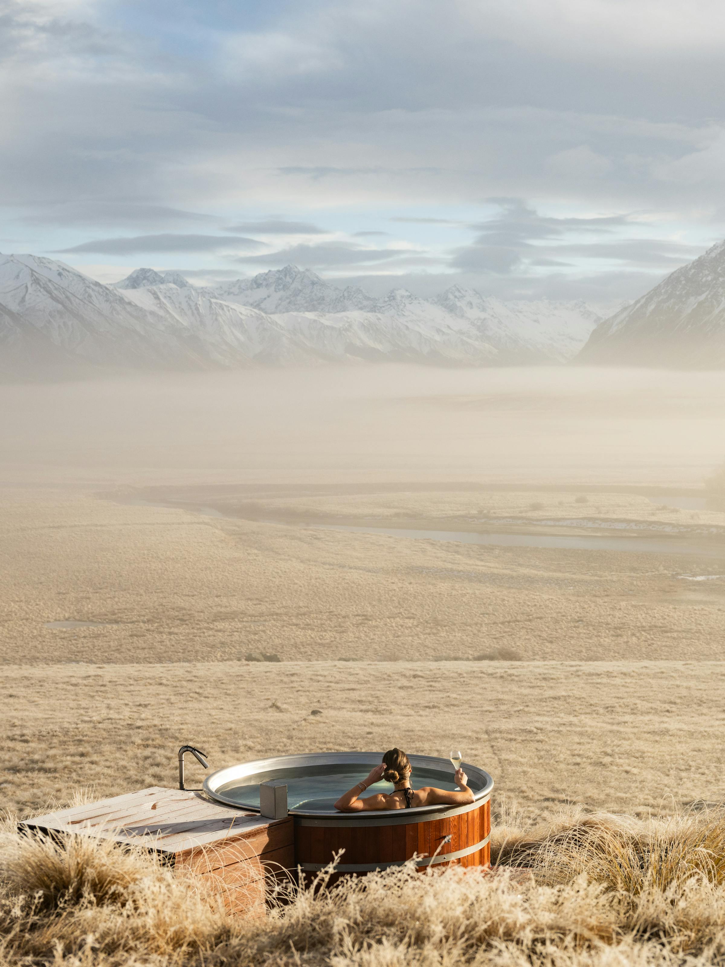 An outdoor hot tub sits in dry tussock grass as a lone guest looks across a misty valley toward rugged peaks.