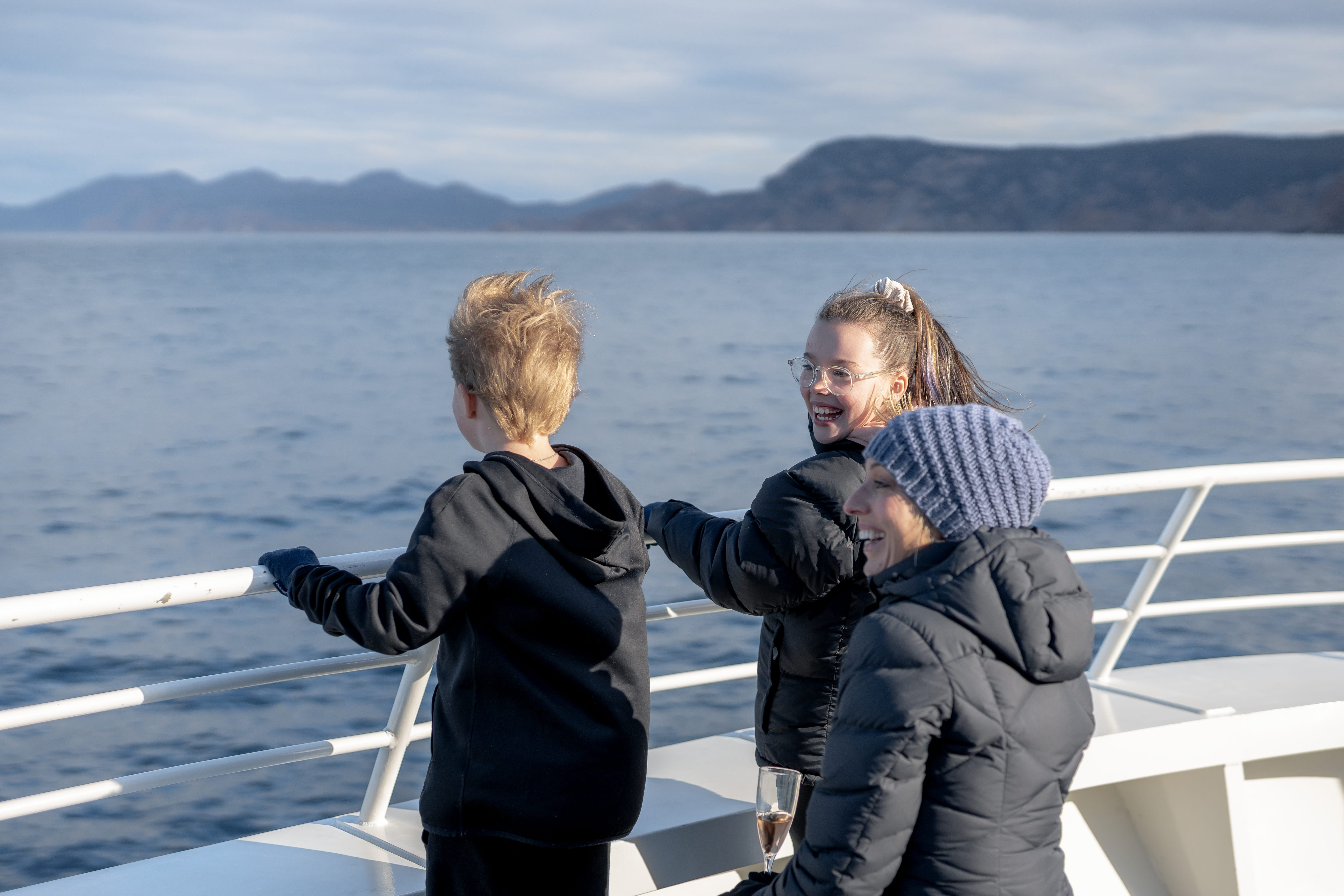 Three passengers in jackets chat on an open boat deck, leaning on the rail with calm sea and hills behind.