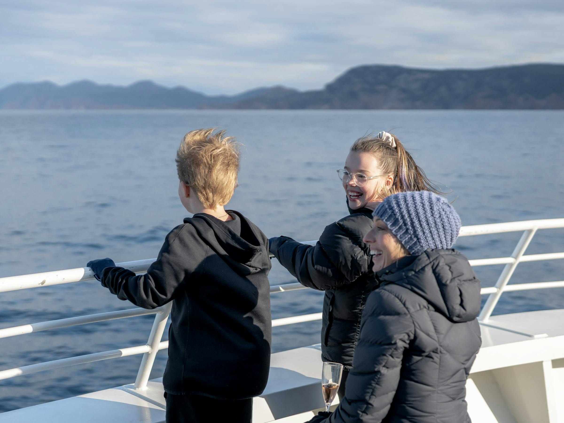 Three passengers in jackets chat on an open boat deck, leaning on the rail with calm sea and hills behind.