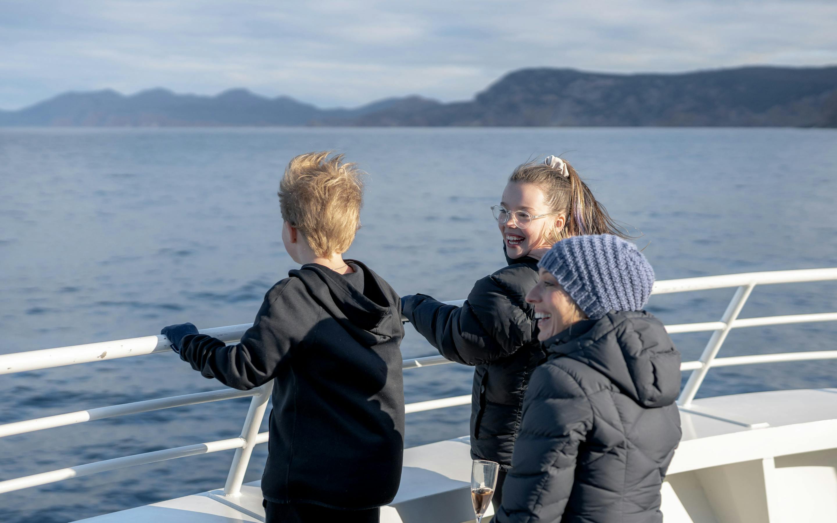 Three passengers in jackets chat on an open boat deck, leaning on the rail with calm sea and hills behind.