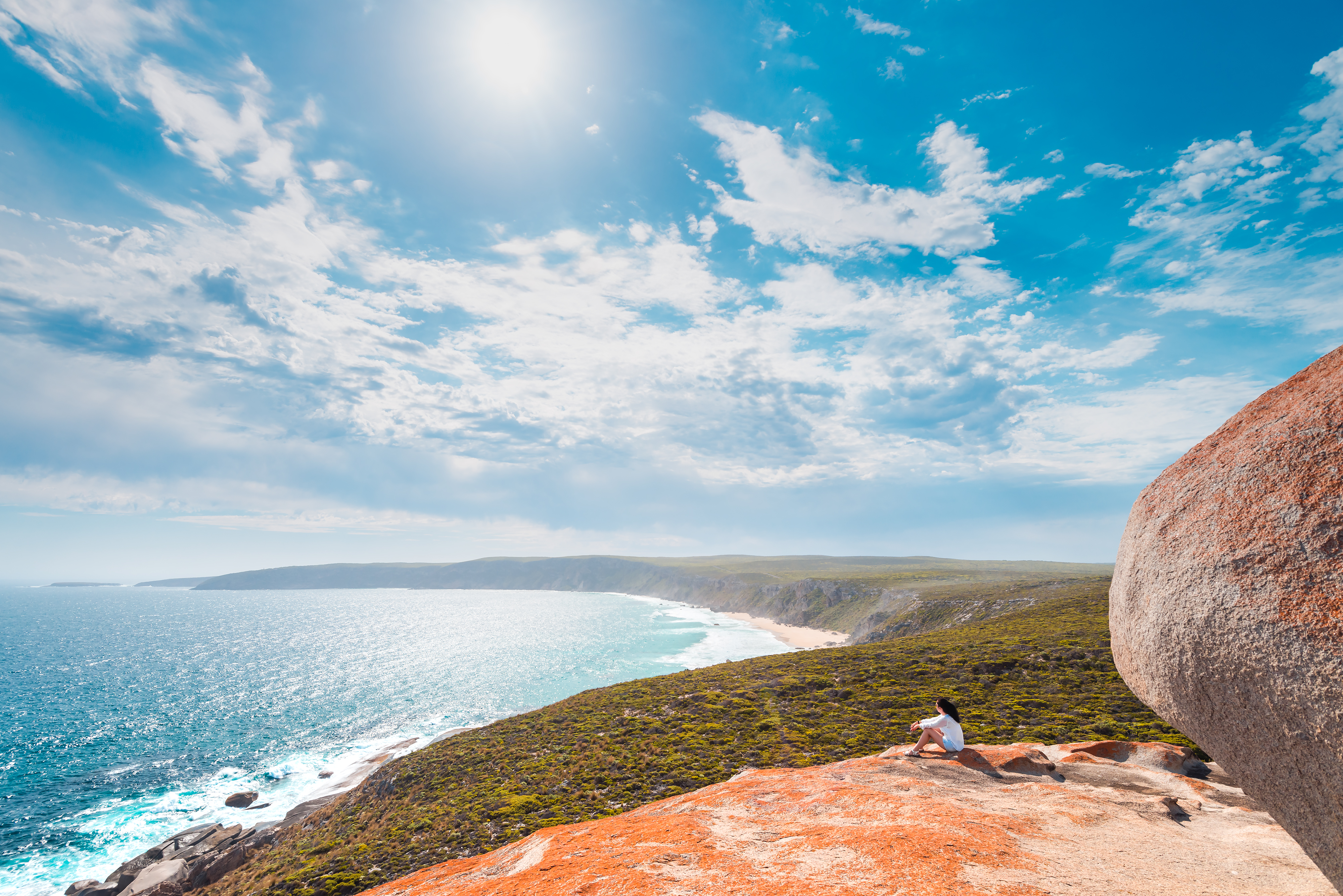 A person sits on a rocky cliff overlooking a bright blue ocean, with sunlit clouds and shoreline stretching below.
