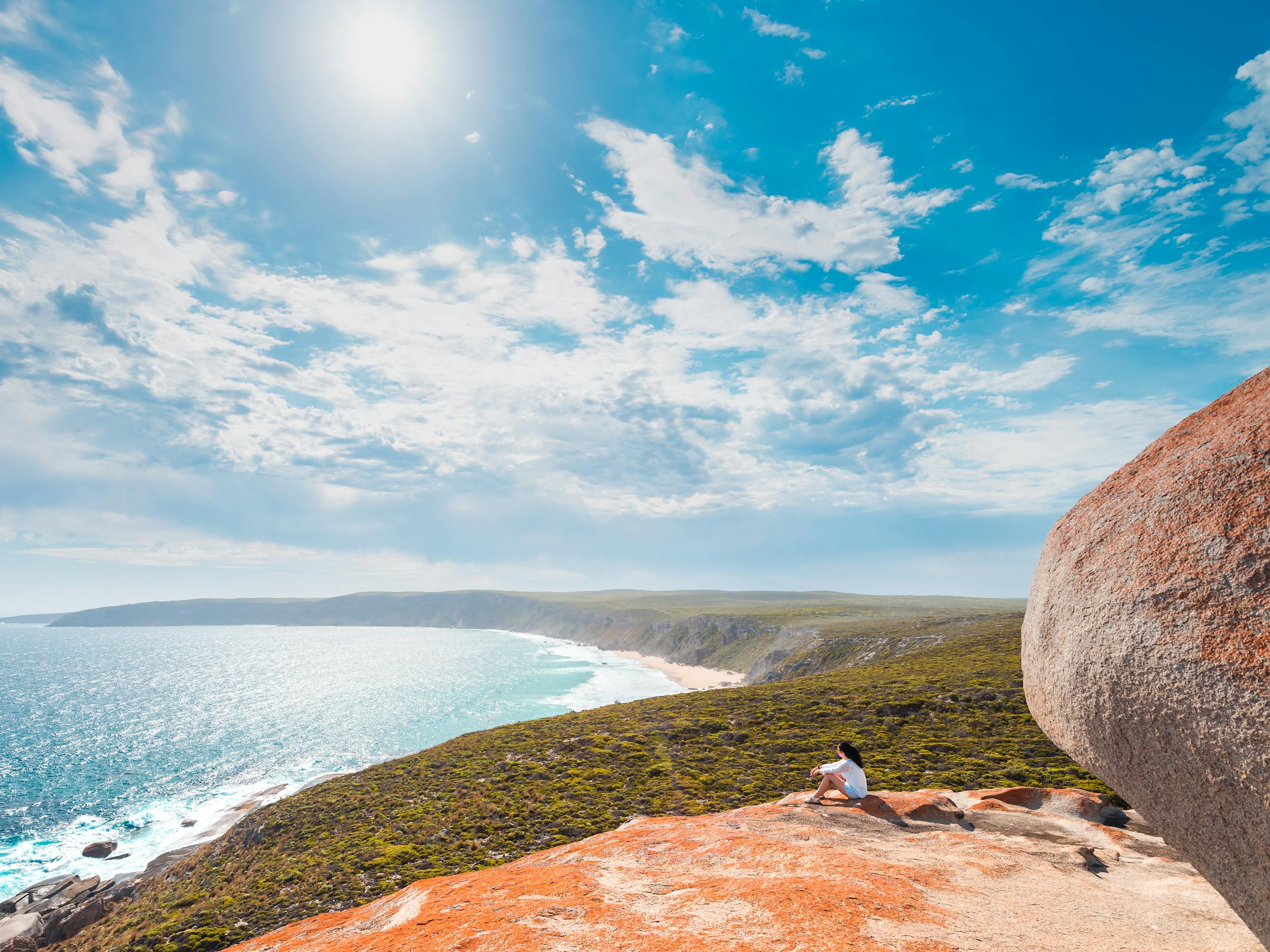 A person sits on a rocky cliff overlooking a bright blue ocean, with sunlit clouds and shoreline stretching below.
