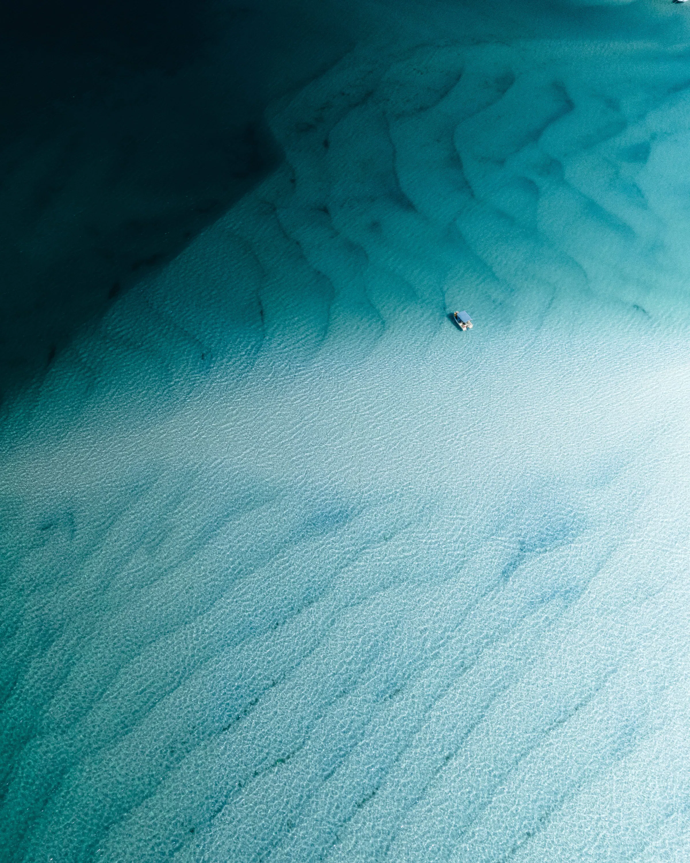 A lone boat floats over pale sandbars in clear turquoise water, with darker reef patches fading into blue.