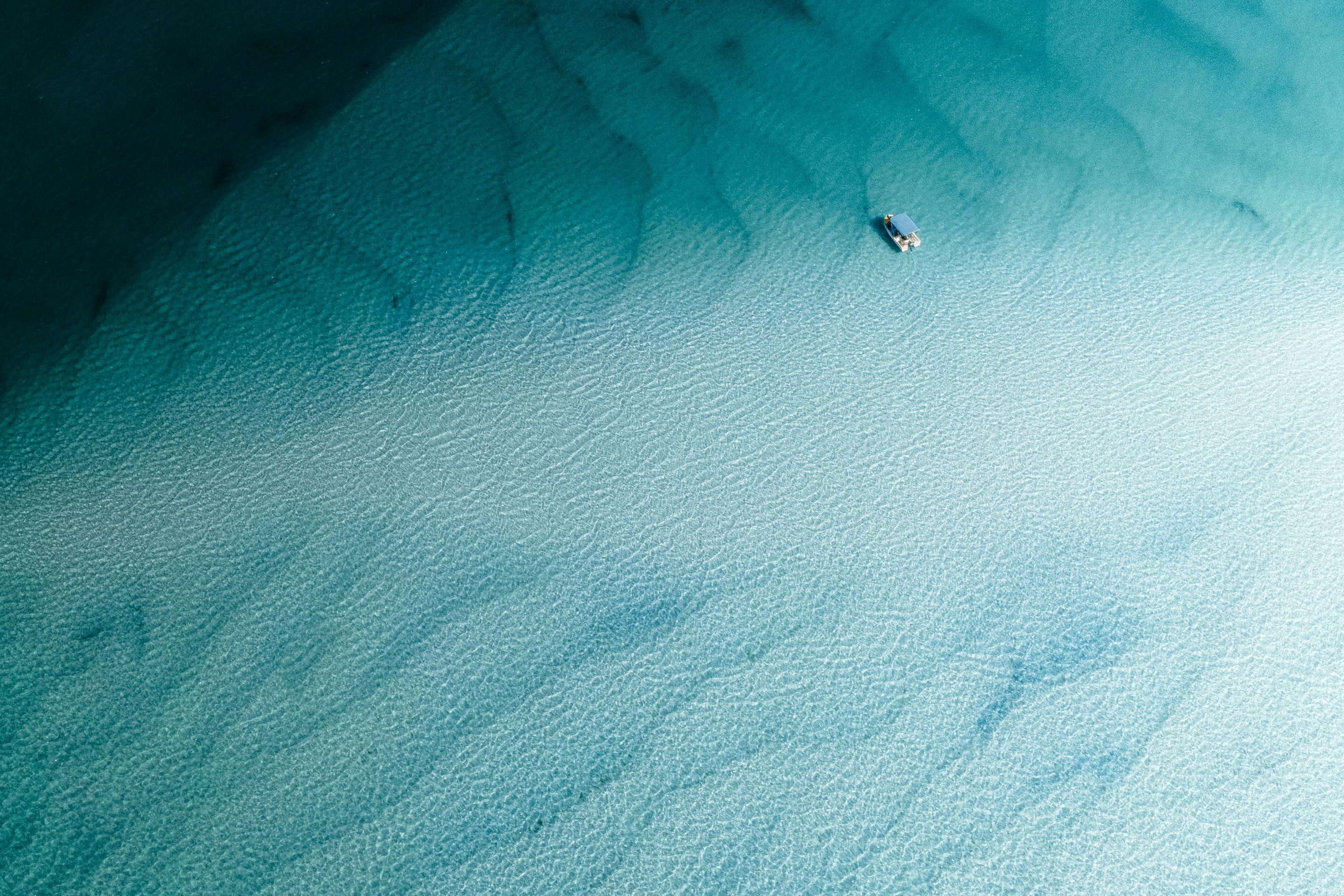 A lone boat floats over pale sandbars in clear turquoise water, with darker reef patches fading into blue.