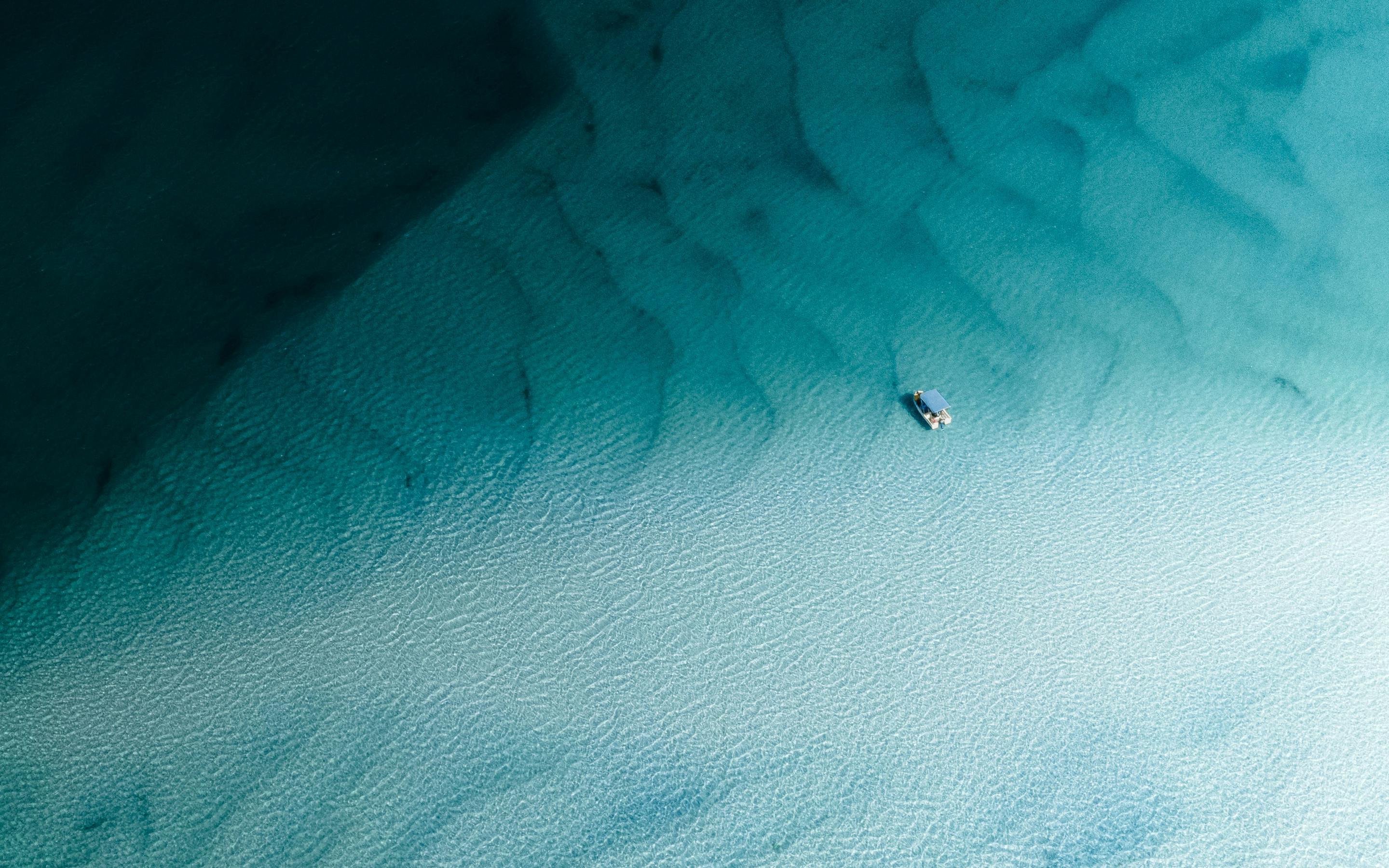 A lone boat floats over pale sandbars in clear turquoise water, with darker reef patches fading into blue.