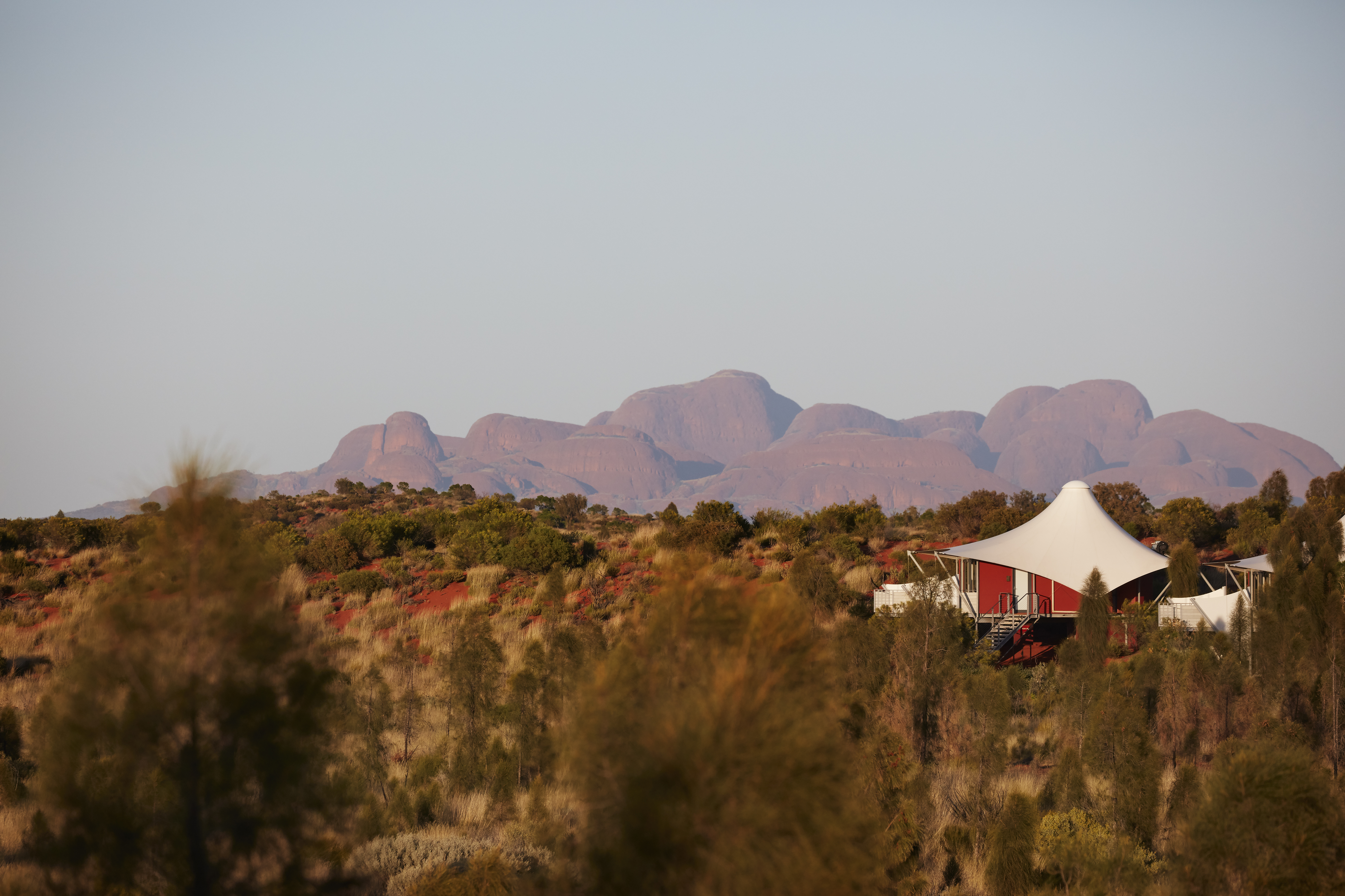 Luxury safari tents sit amid desert scrub, with Kata Tjuta rising in hazy light beyond the dunes at dusk.