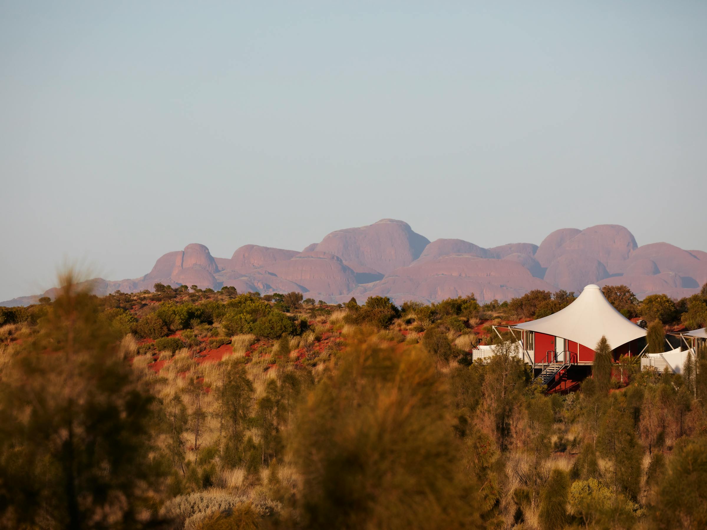 Luxury safari tents sit amid desert scrub, with Kata Tjuta rising in hazy light beyond the dunes at dusk.