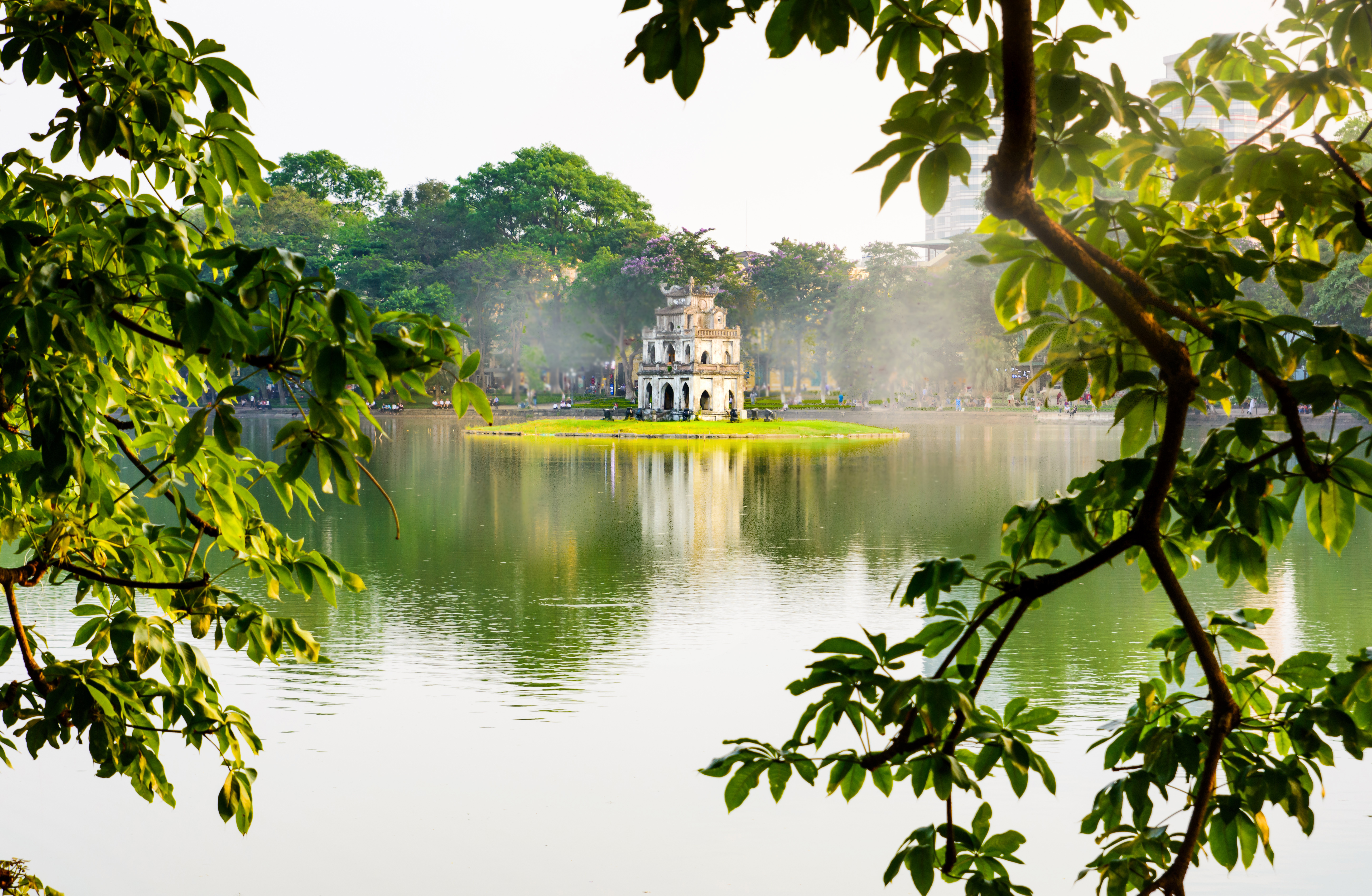 Turtle Tower rises from Hoan Kiem Lake, framed by leafy branches and calm water reflecting soft morning light.