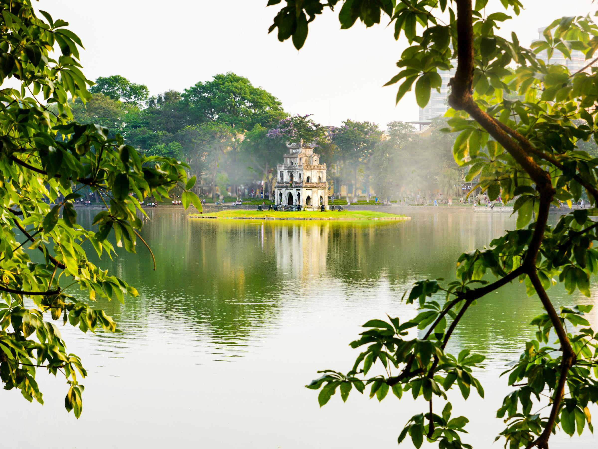 Turtle Tower rises from Hoan Kiem Lake, framed by leafy branches and calm water reflecting soft morning light.