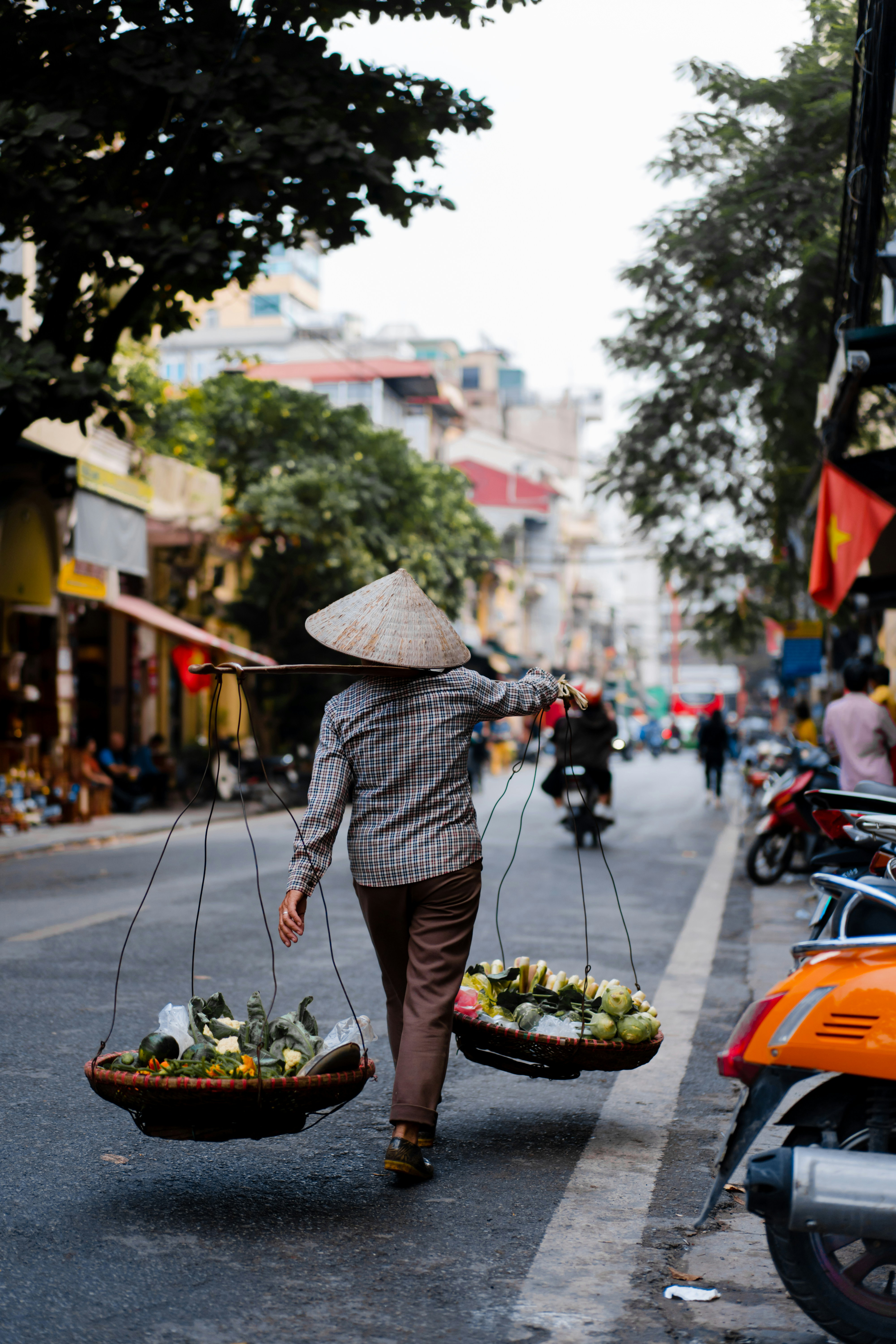 A vendor carries a yoke with baskets of produce along a Hanoi street as motorbikes and shops blur behind.
