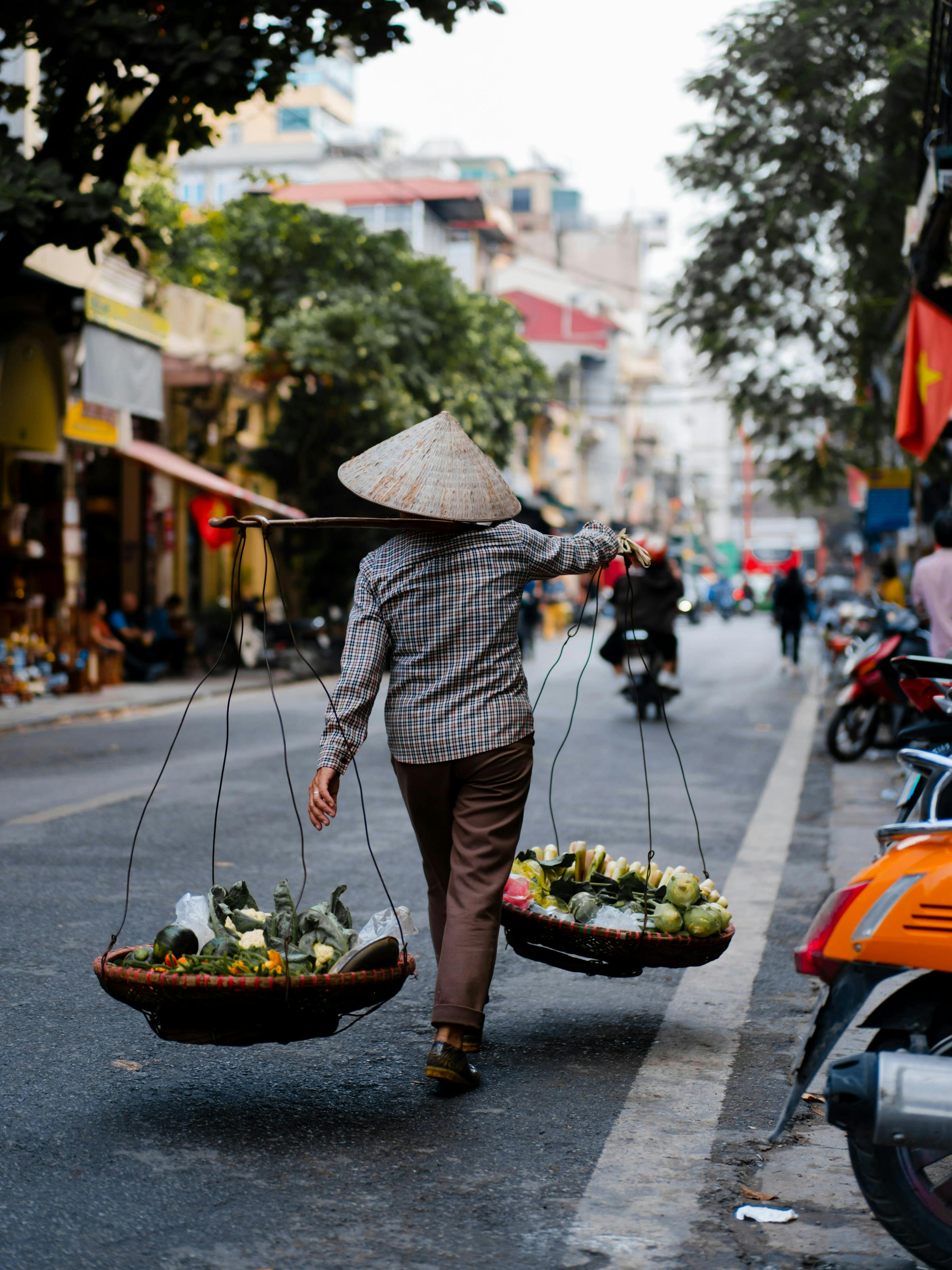 A vendor carries a yoke with baskets of produce along a Hanoi street as motorbikes and shops blur behind.