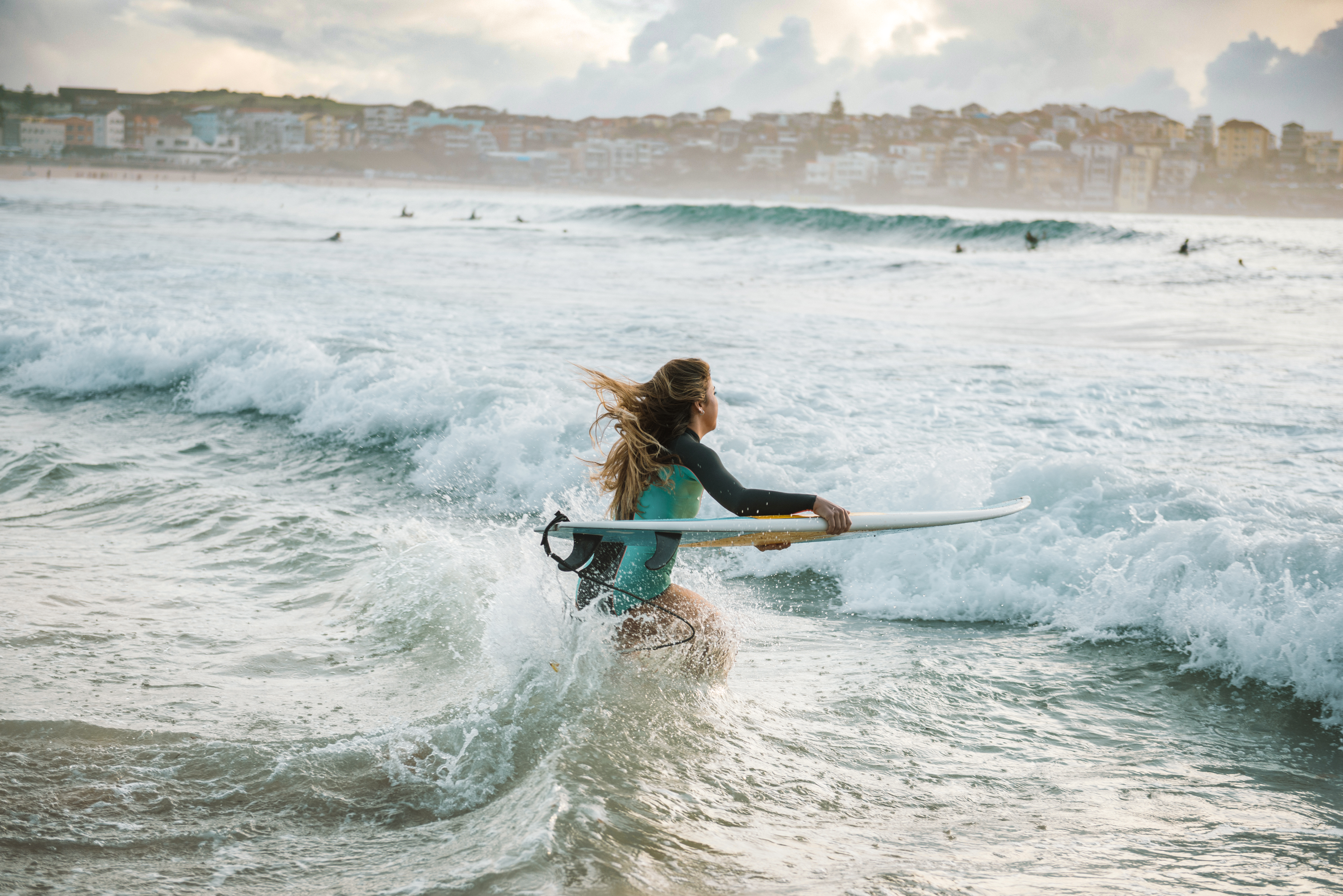 Surfer wades into breaking waves carrying a longboard, with other surfers and a hazy coastline in the distance.