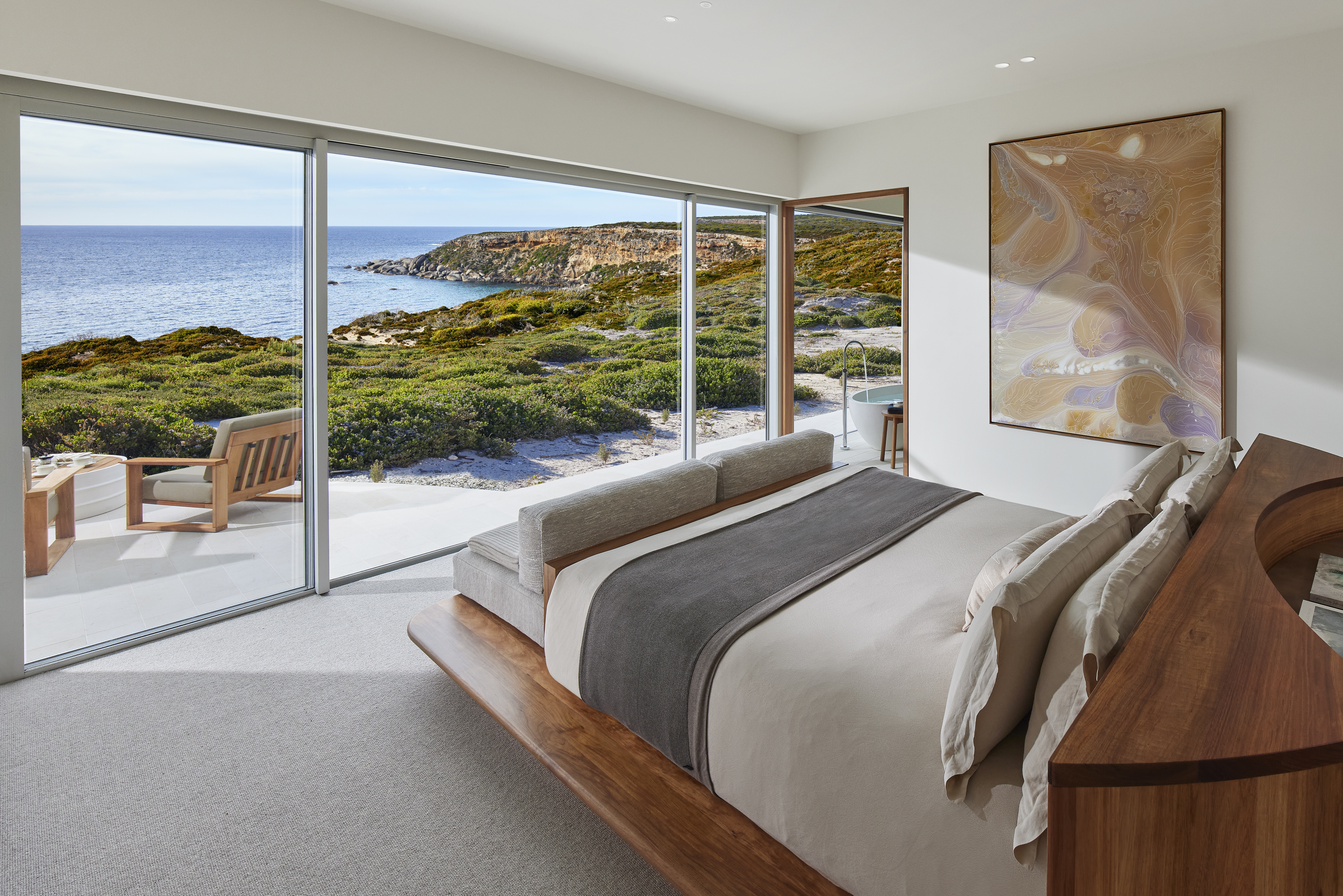 A modern bedroom opens to an ocean-view terrace, with a neatly made bed and wide glass doors to the sea beyond.