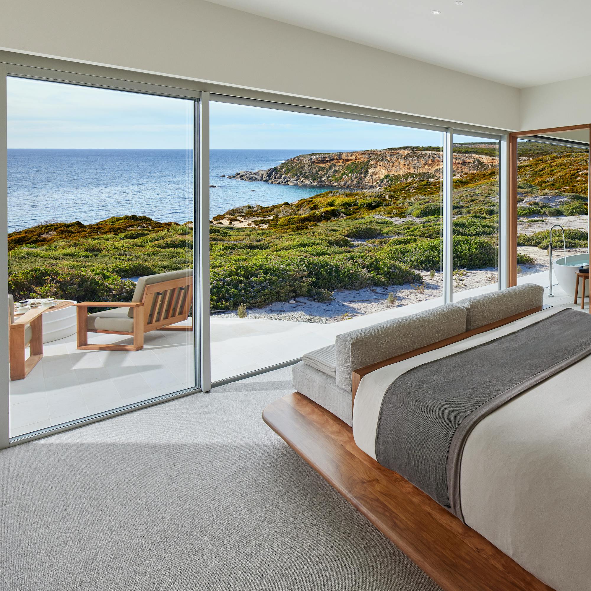 A modern bedroom opens to an ocean-view terrace, with a neatly made bed and wide glass doors to the sea beyond.