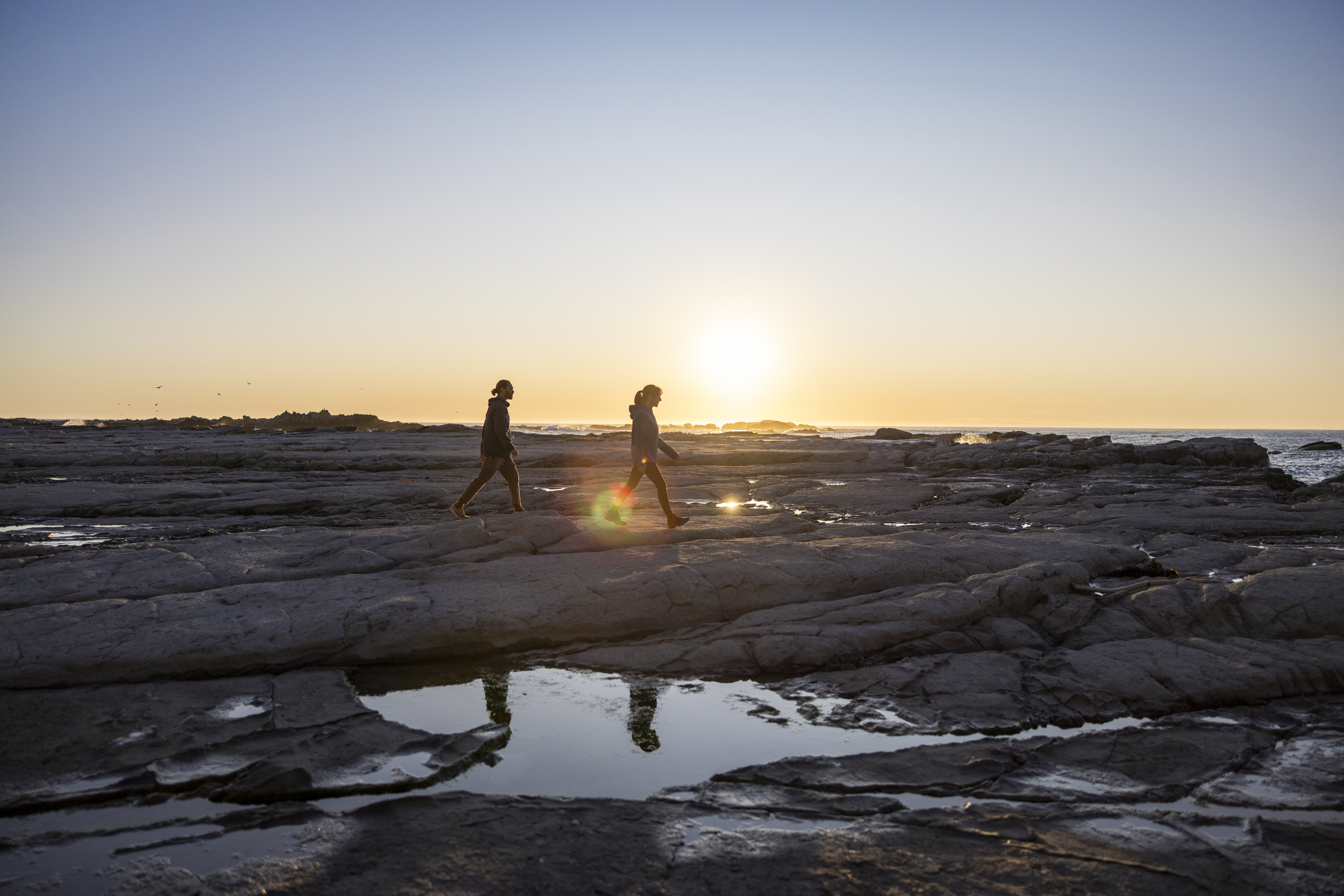Two people walk across rocky tide pools at sunset near Kaikoura, with reflections glowing in shallow water.