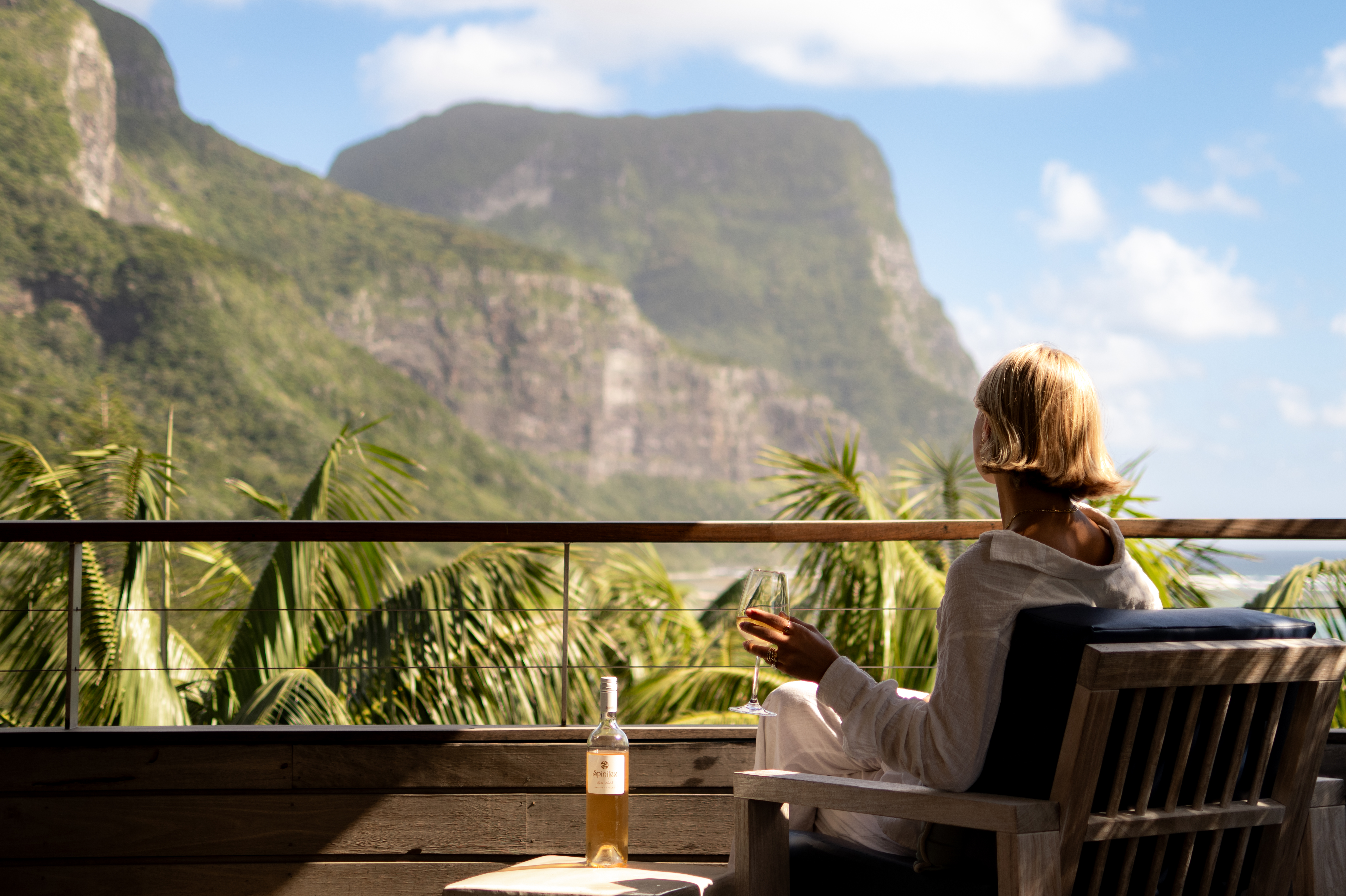 A guest relaxes on a lodge veranda with breakfast, facing steep green mountains rising above palm trees and clouds.