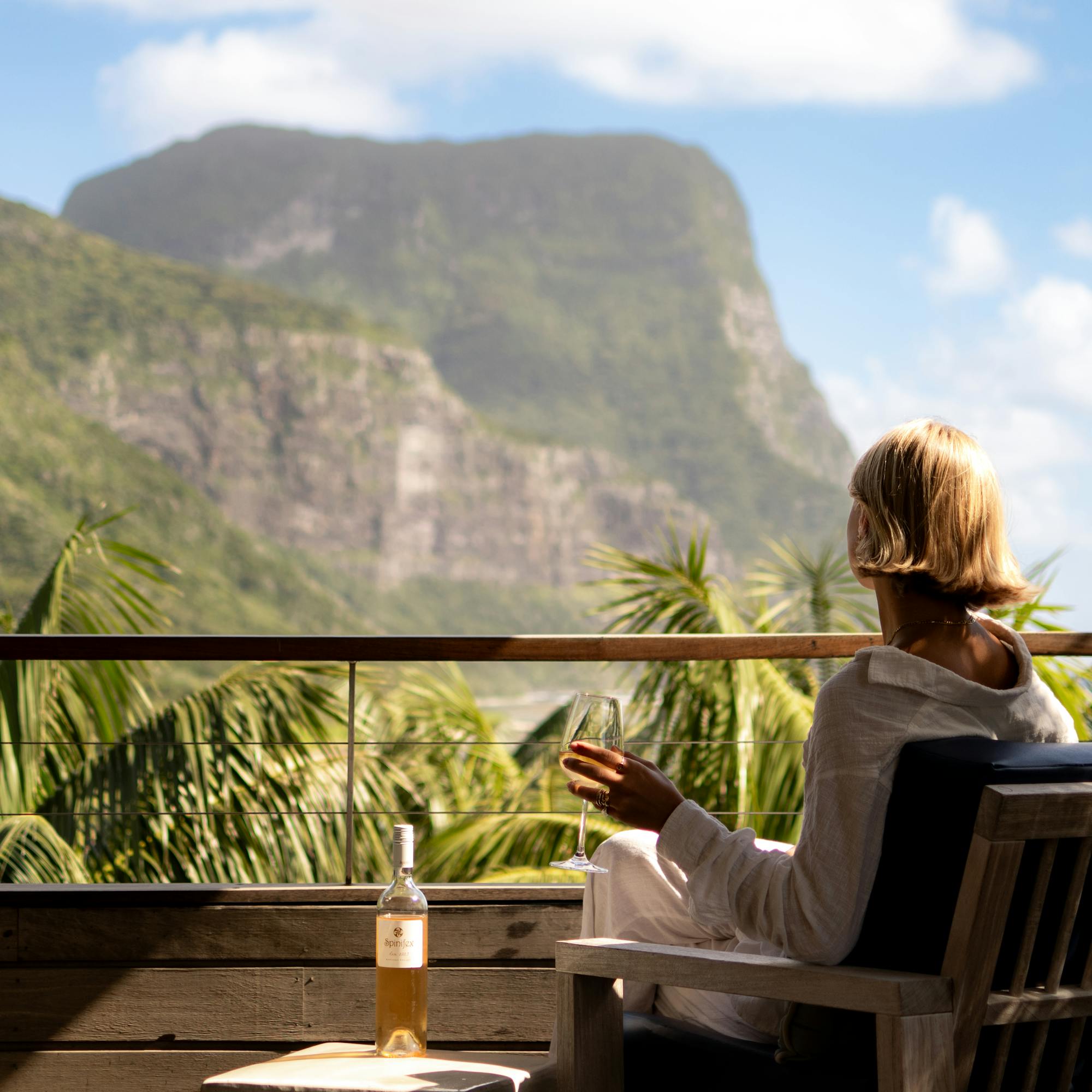 A guest relaxes on a lodge veranda with breakfast, facing steep green mountains rising above palm trees and clouds.