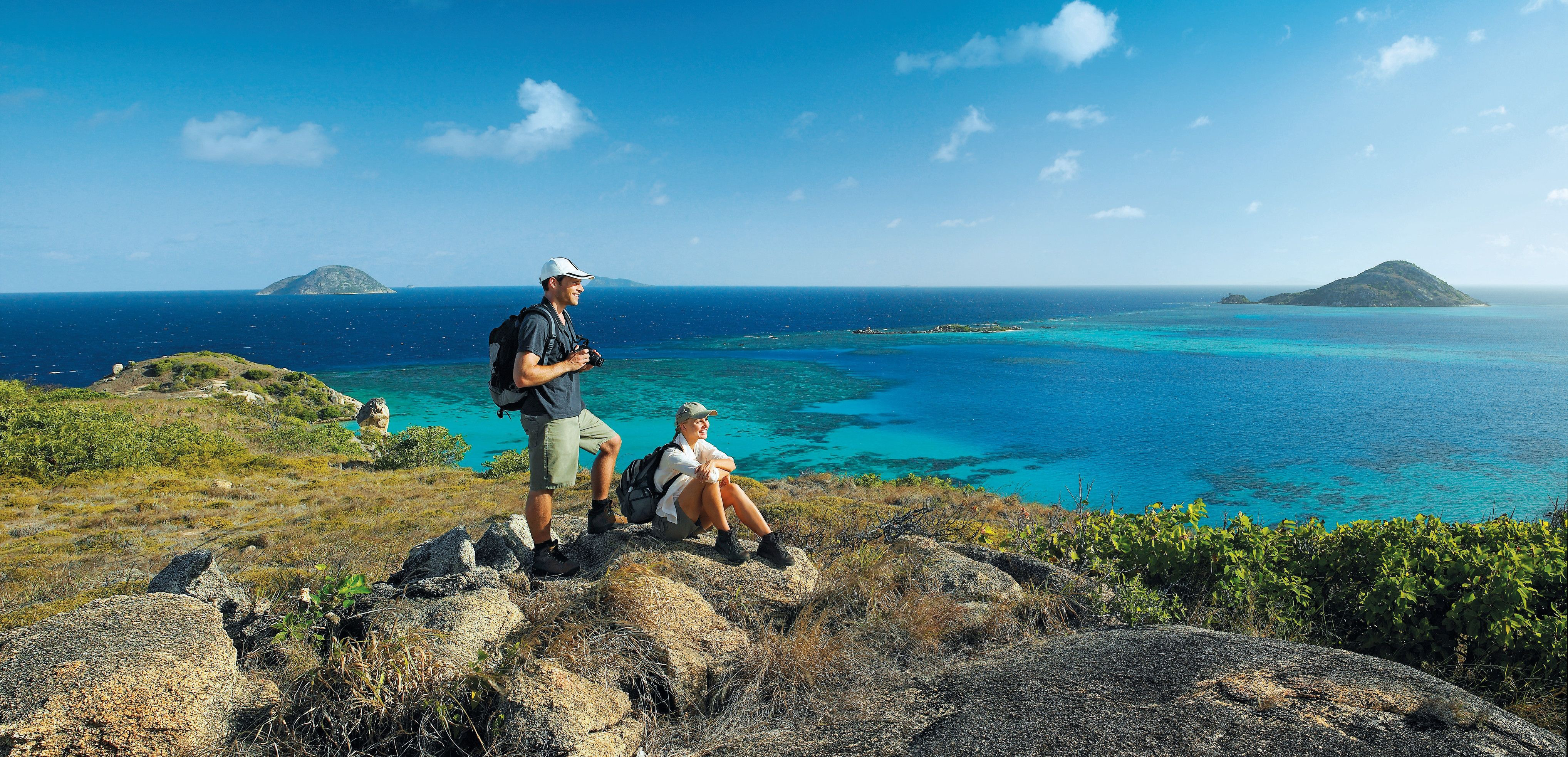 Two hikers rest on a rocky ridge, looking across a lagoon of vivid blues toward small islands under a clear sky.