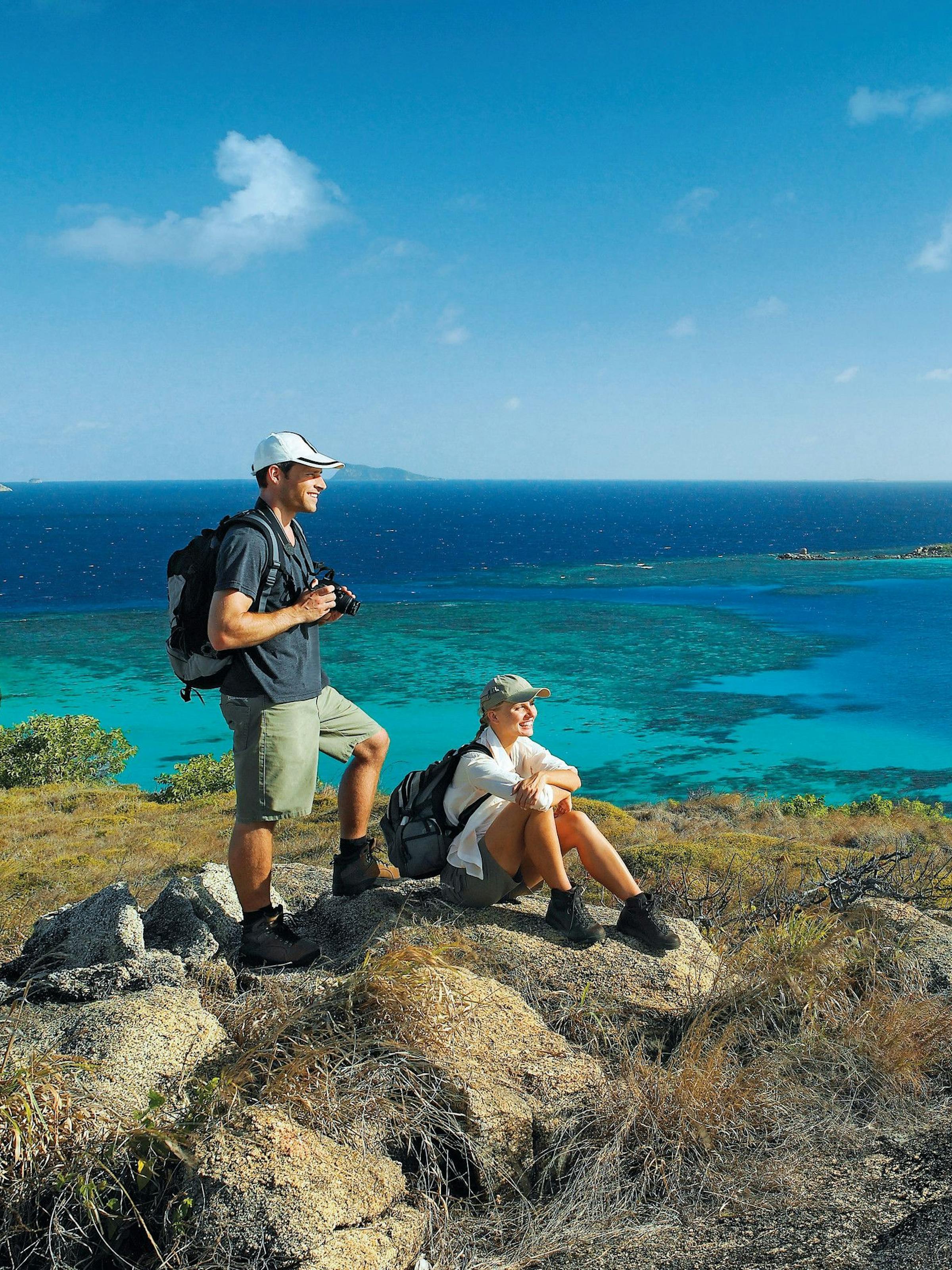 Two hikers rest on a rocky ridge, looking across a lagoon of vivid blues toward small islands under a clear sky.