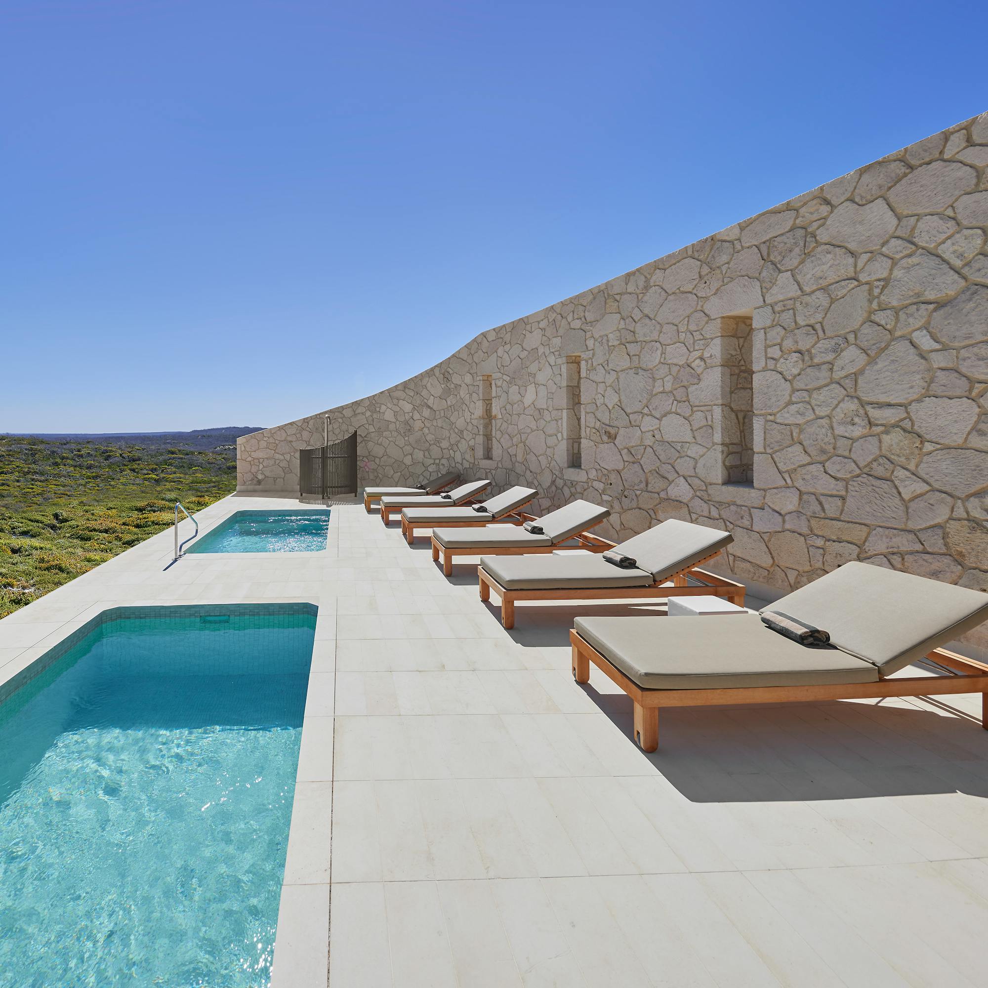 Sunlit lap pool and loungers run beside a stone wall, overlooking rolling scrubland and distant ocean horizon.