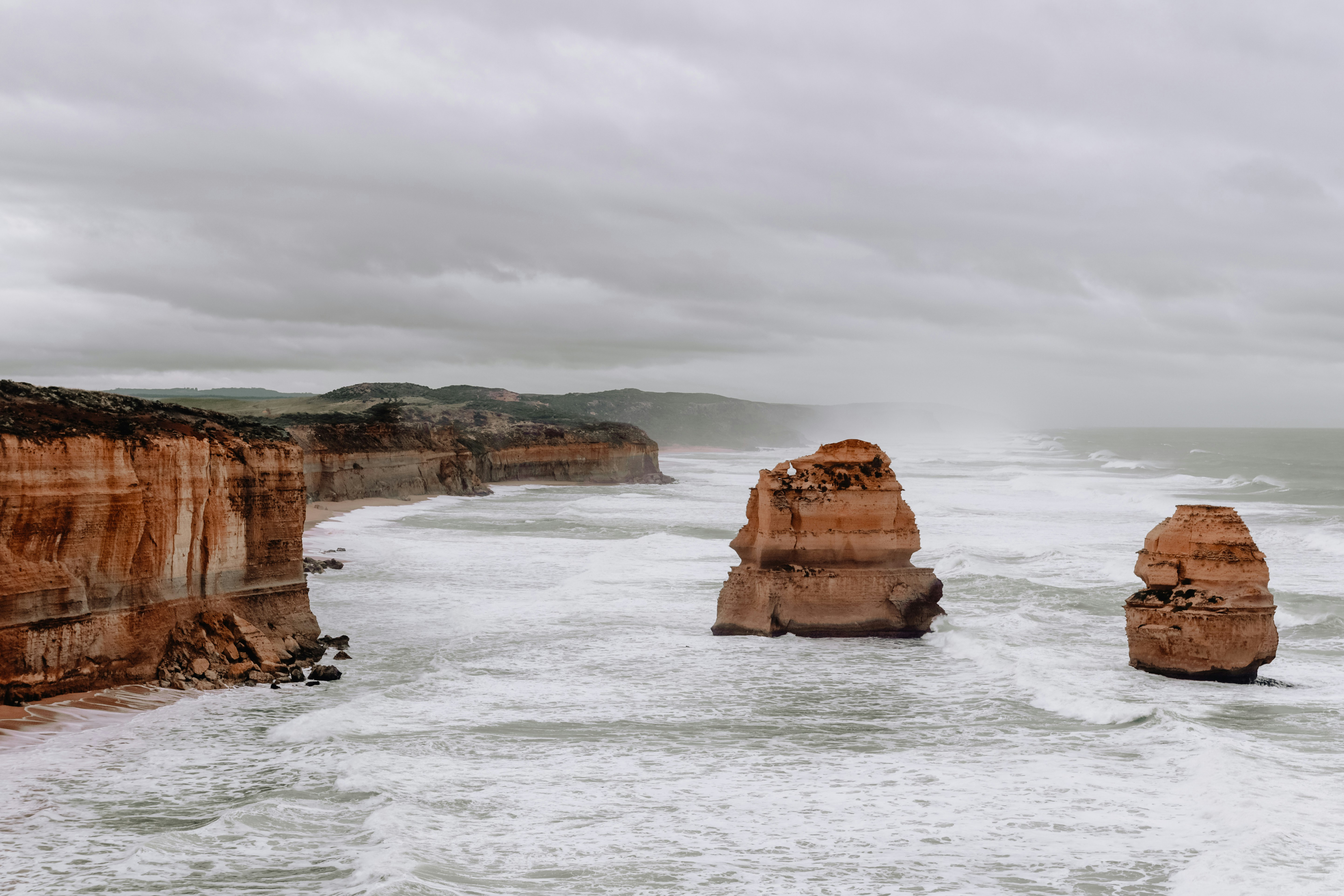 Sandstone cliffs and offshore sea stacks rise from choppy grey water, with low clouds hanging over the coastline.