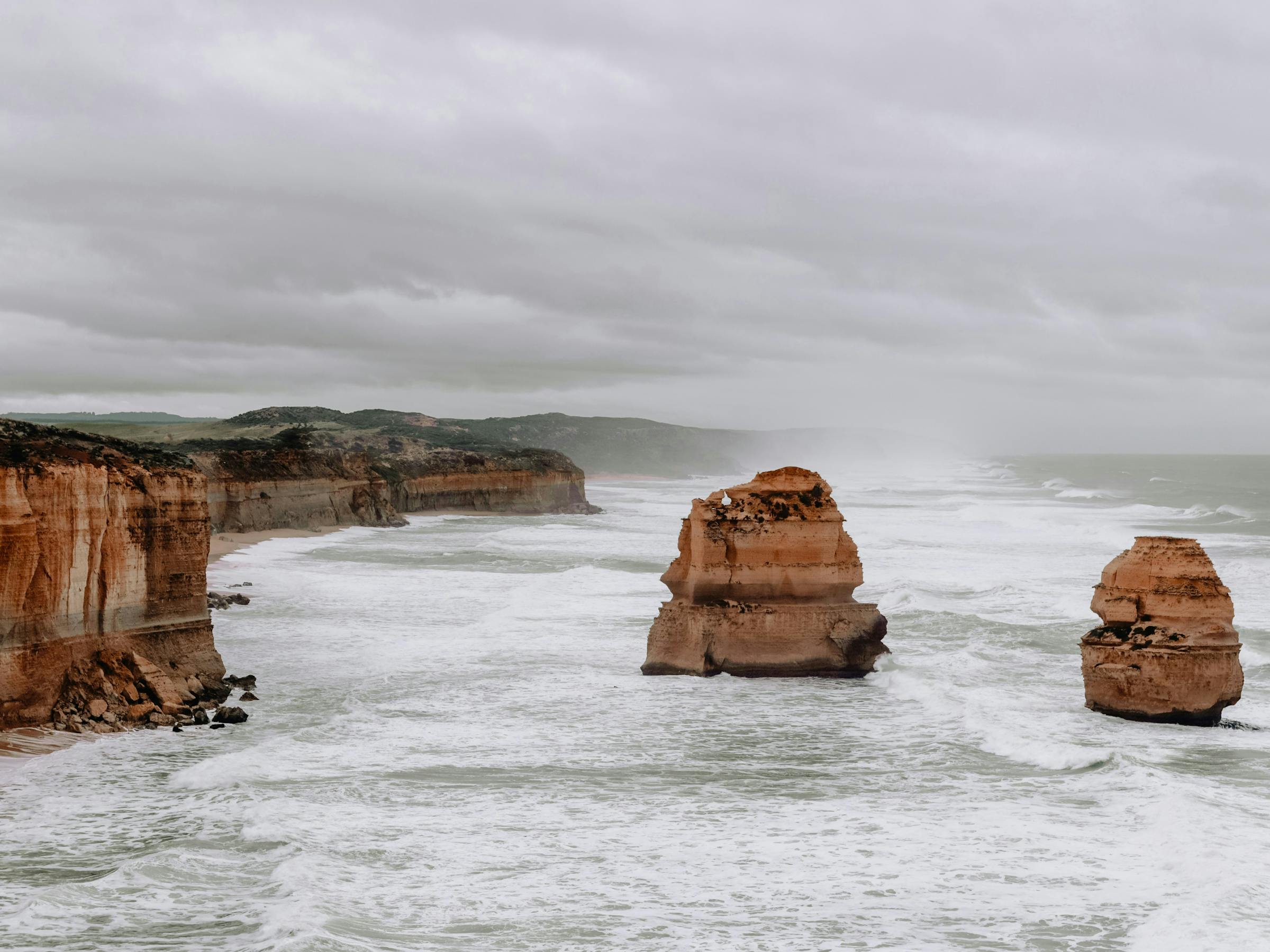 Sandstone cliffs and offshore sea stacks rise from choppy grey water, with low clouds hanging over the coastline.