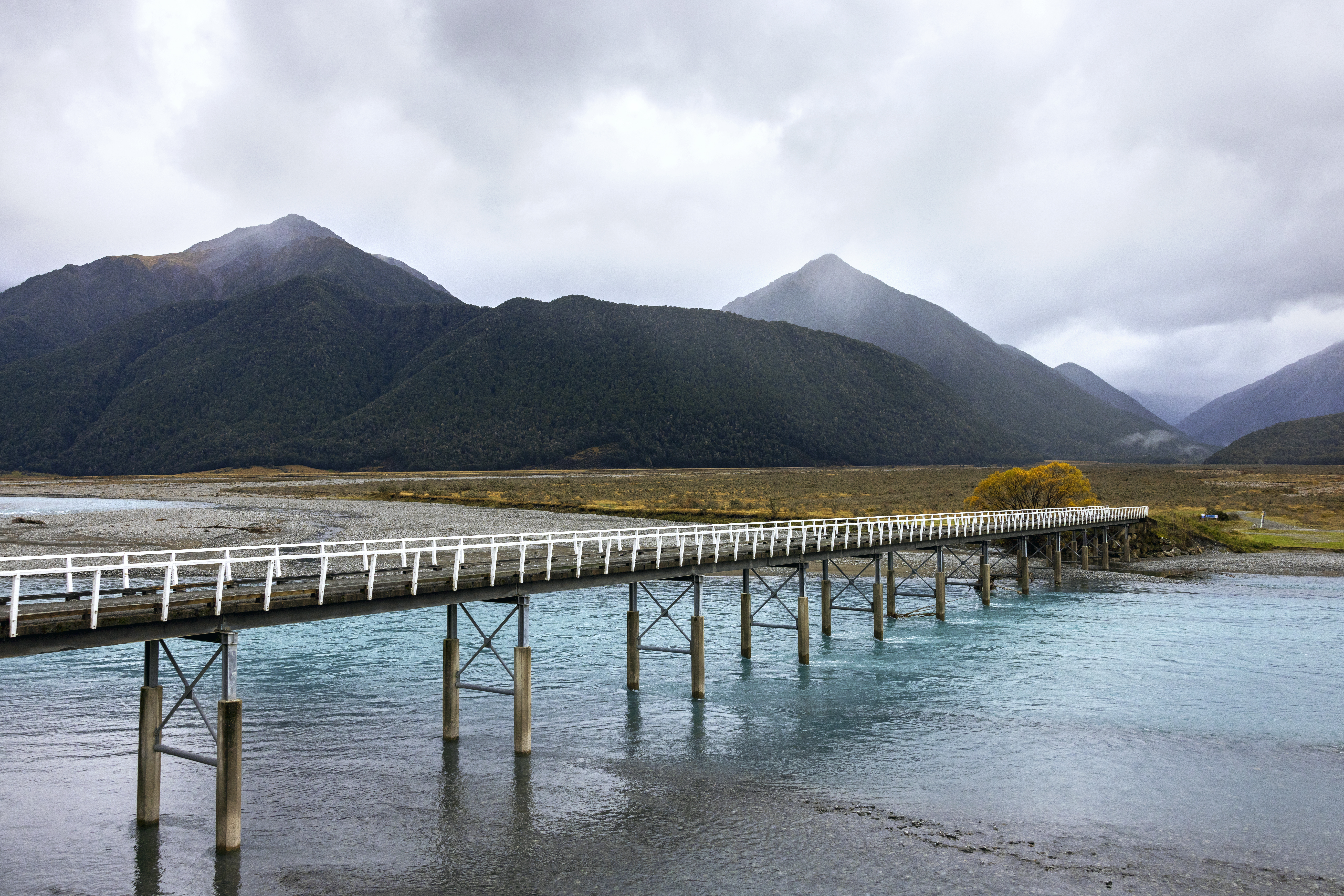 Long wooden pier extends over clear lake water toward dark mountains, with golden grasses and misty light beyond.
